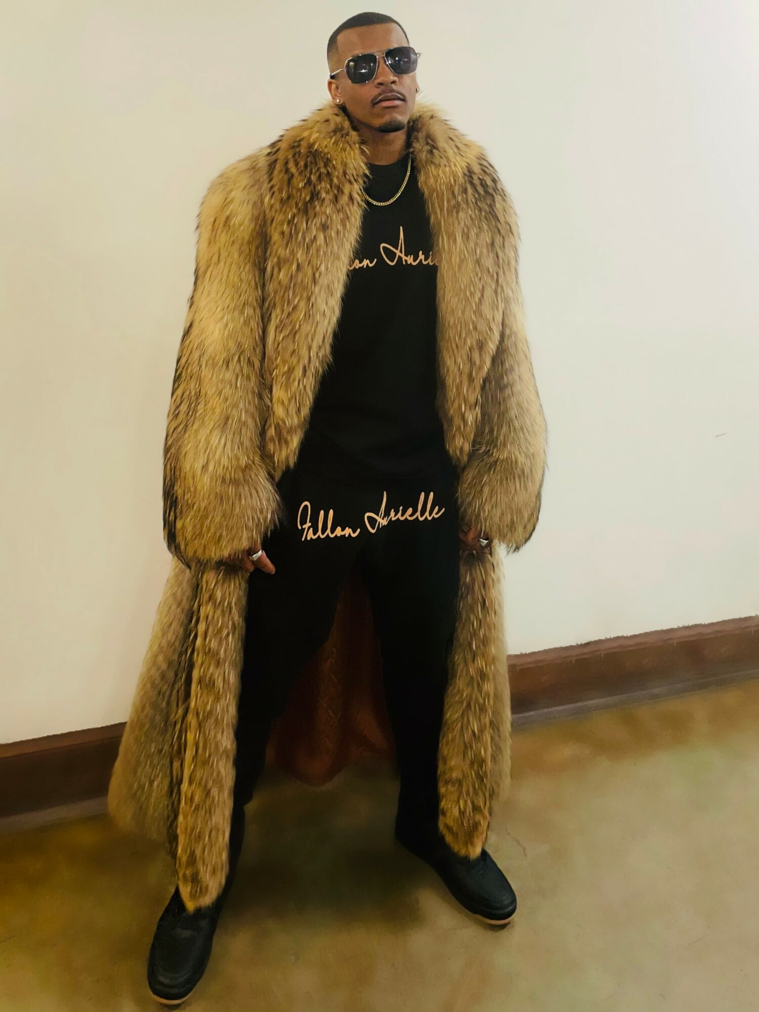 Man in sunglasses wearing a long fur coat, black shirt, black pants, and black shoes, standing against a plain wall.