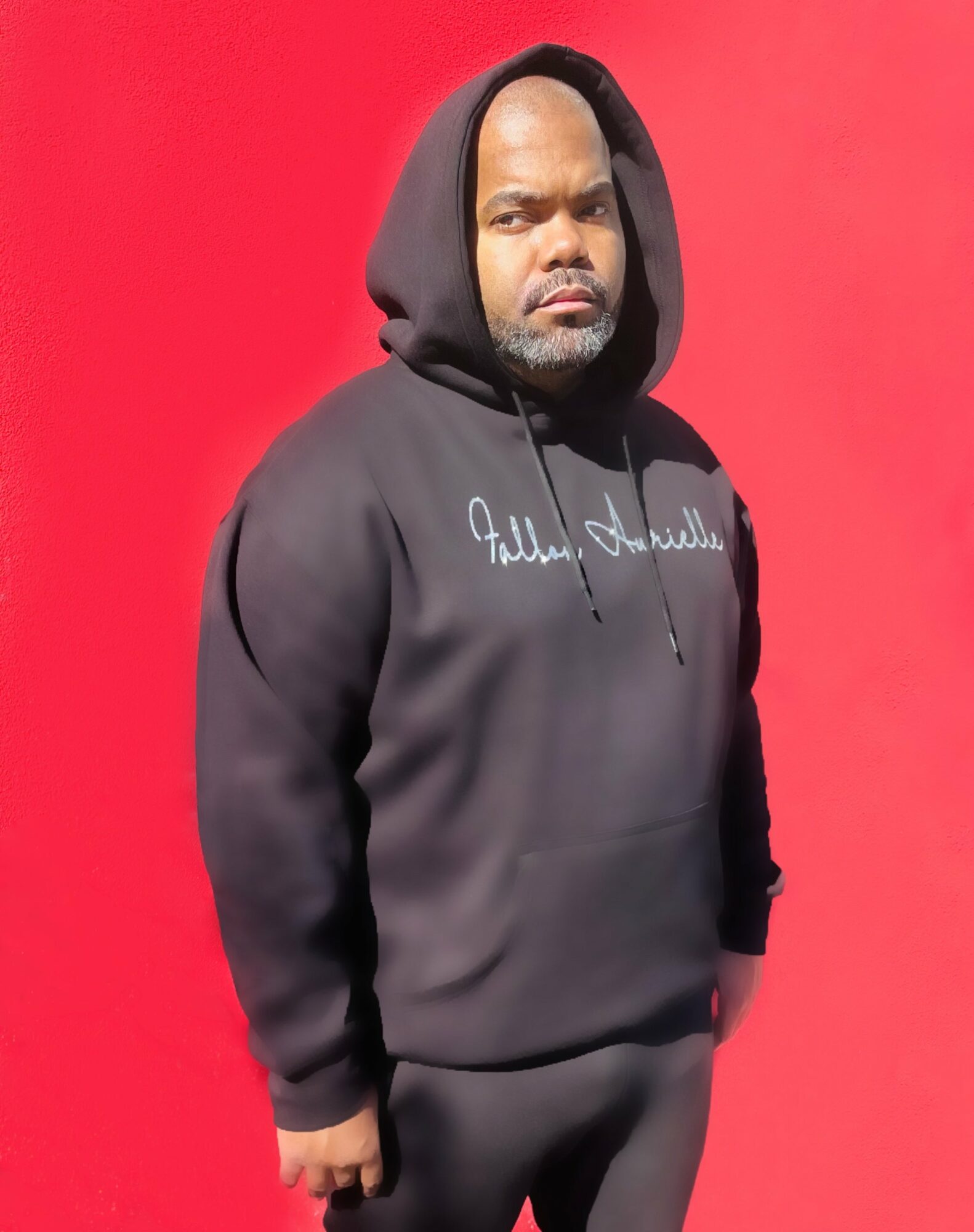 Man wearing a black hoodie with white text, standing against a red background, looking at the camera.
