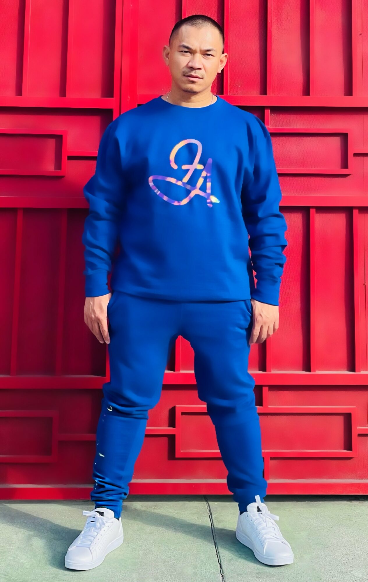 Young man in blue sweatshirt and sweatpants standing in front of a red wooden fence.