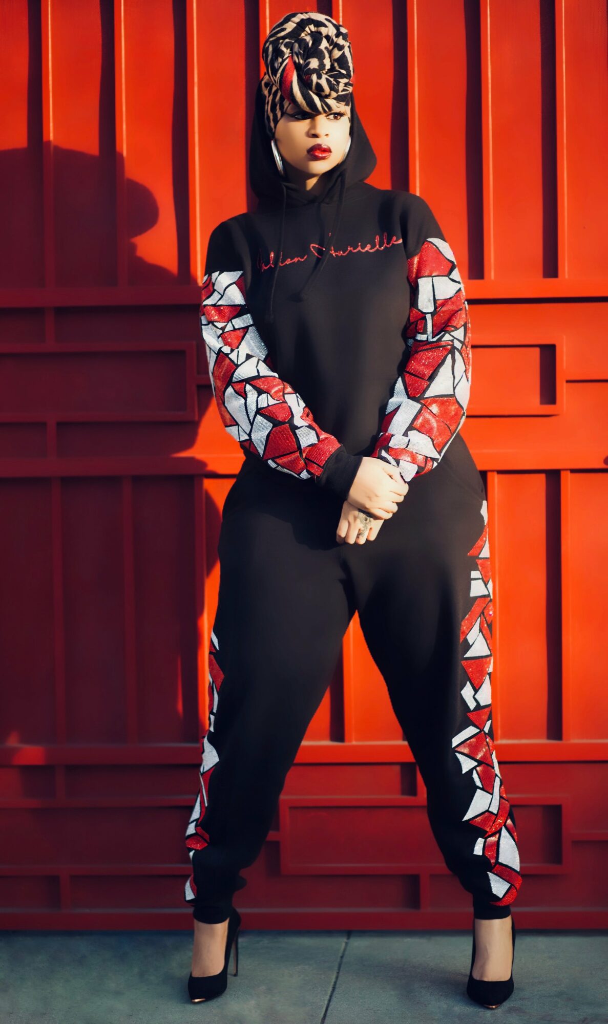 Woman in black and red patterned tracksuit with high heels, standing against a red wooden wall.