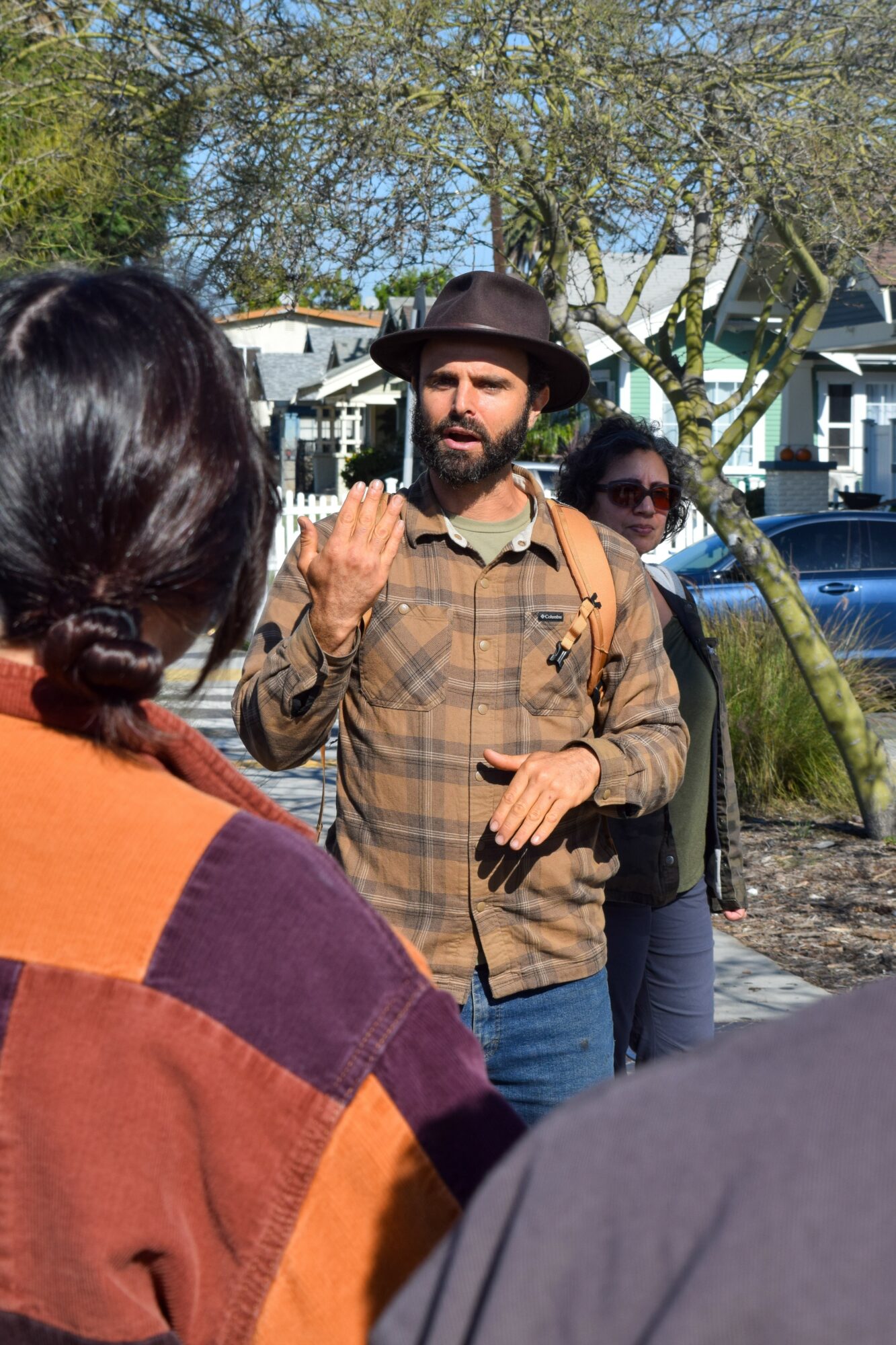 Man with beard wearing a hat and plaid shirt gesturing outdoors with two women nearby.