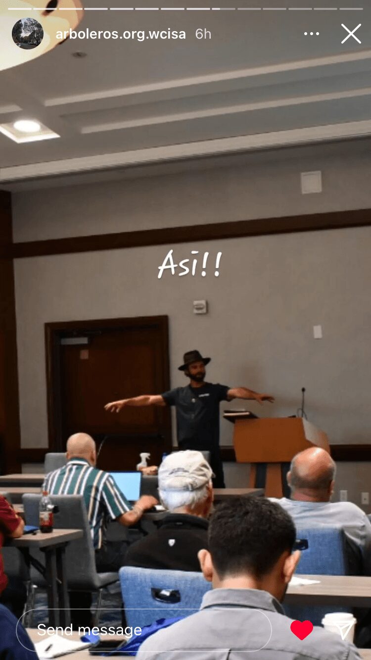 Person standing in front of seated audience, gesturing with arms outstretched in a room with high ceiling and wooden accents.