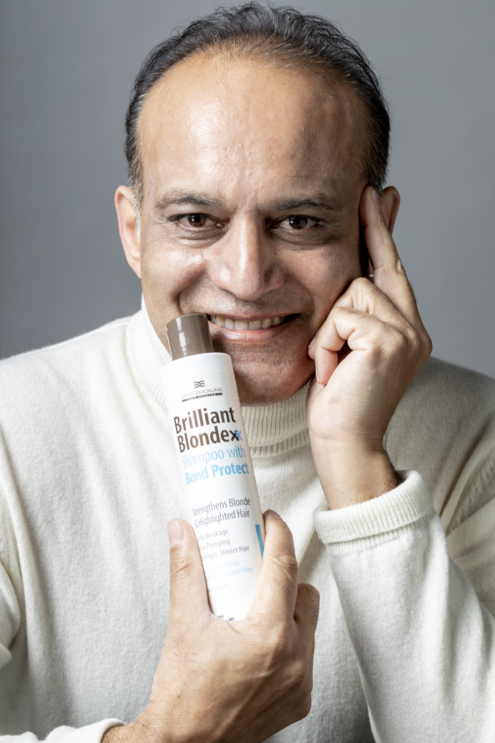 Man smiling, holding a bottle of Brilliant Blondex shampoo near face, touching temple with index finger, gray background.