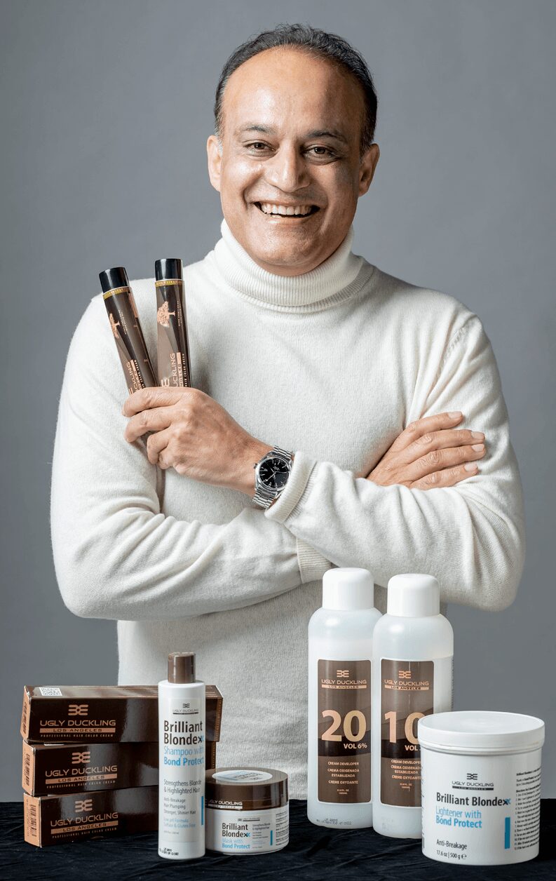 Smiling man holding two bottles, surrounded by skincare products on table, wearing white turtleneck and watch, gray background.