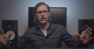 Person with glasses and long hair speaking with hands raised in a room with speakers and a monitor.