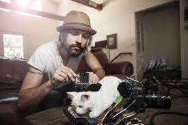 Man with a hat and beard taking a photo of a white dog with black spots on a couch.