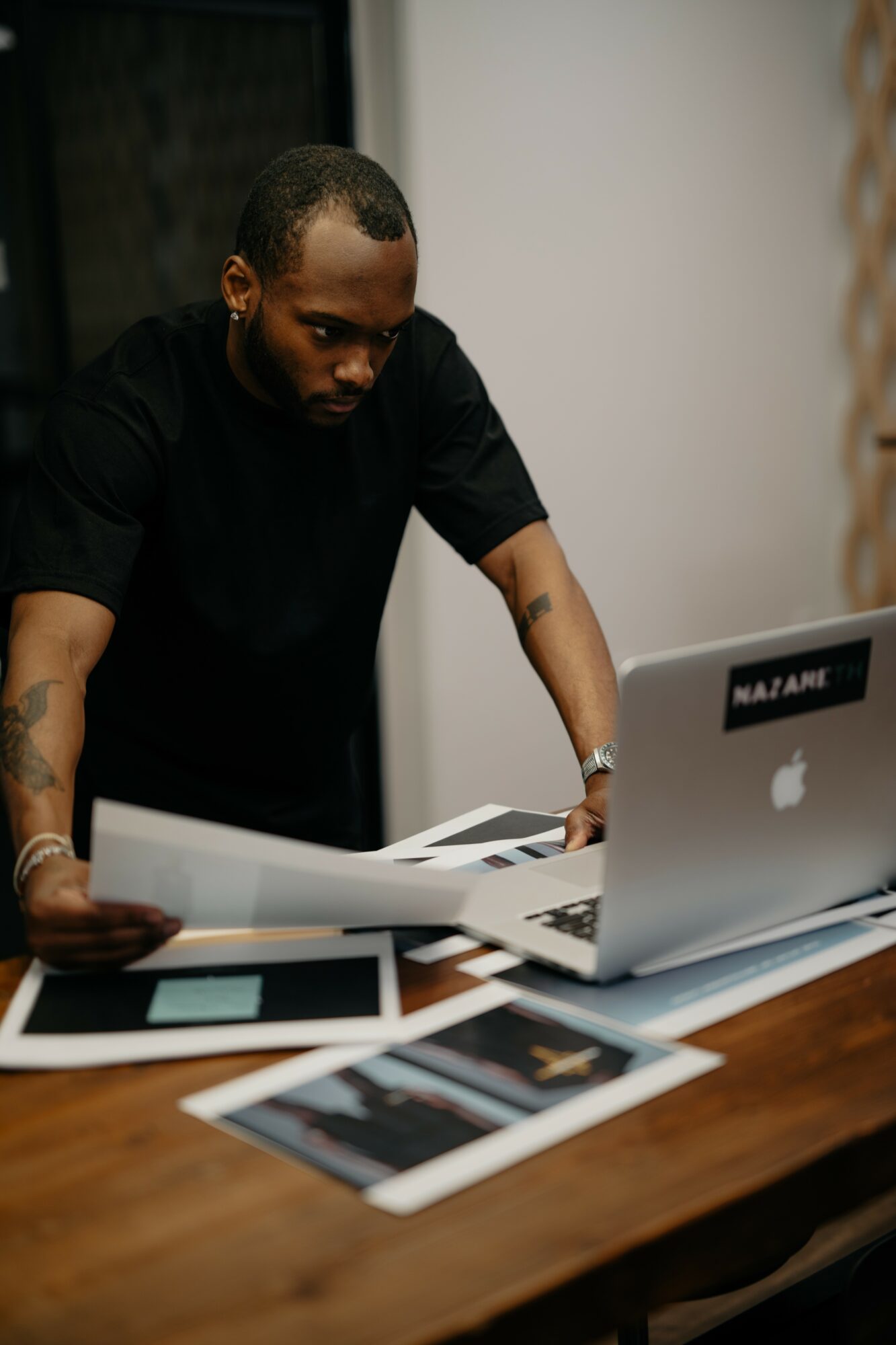 Man in black shirt leaning over a desk with papers and a laptop.
