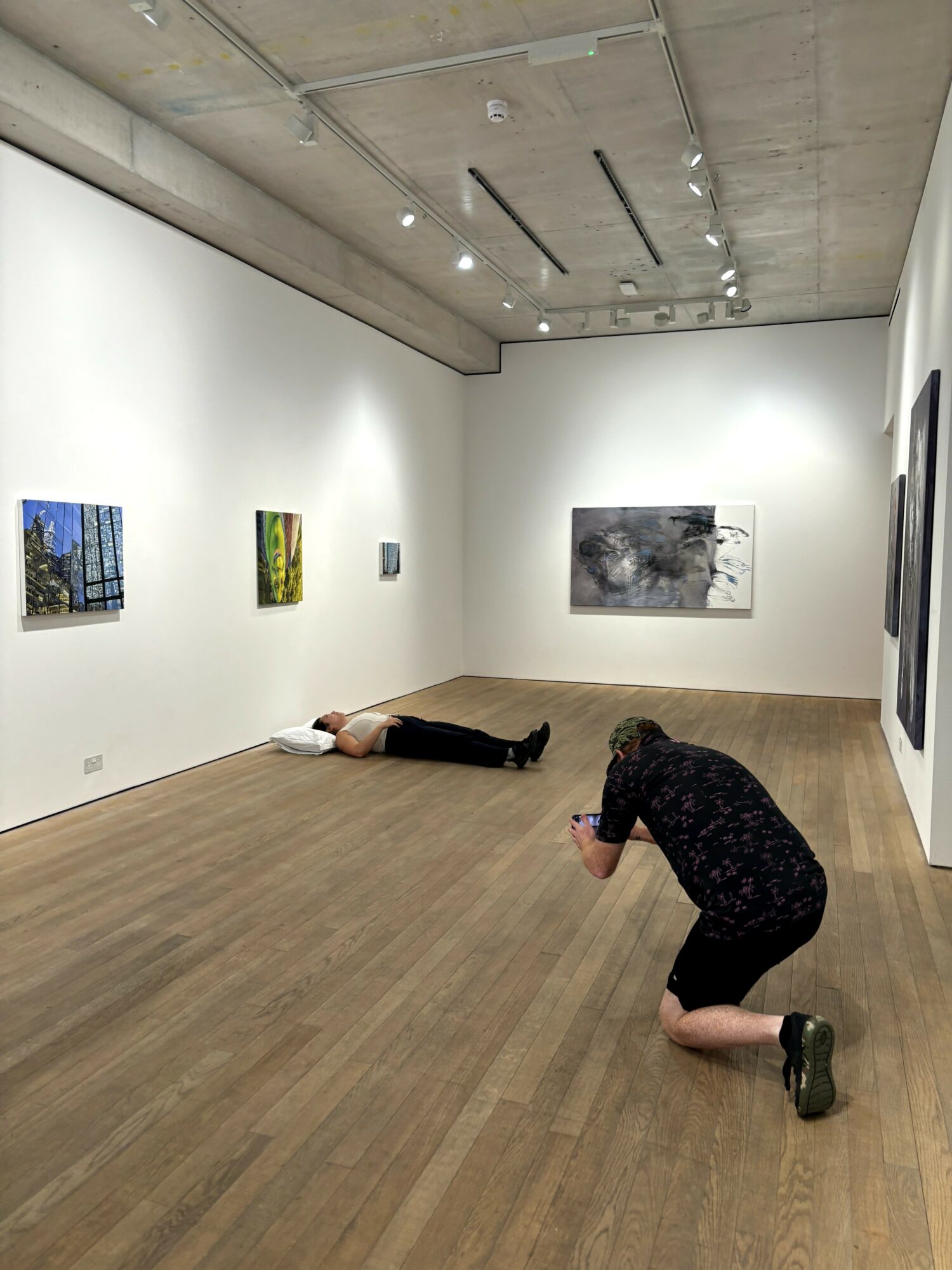 Person kneeling on the floor taking a photo in an art gallery with paintings on white walls and wooden floor.