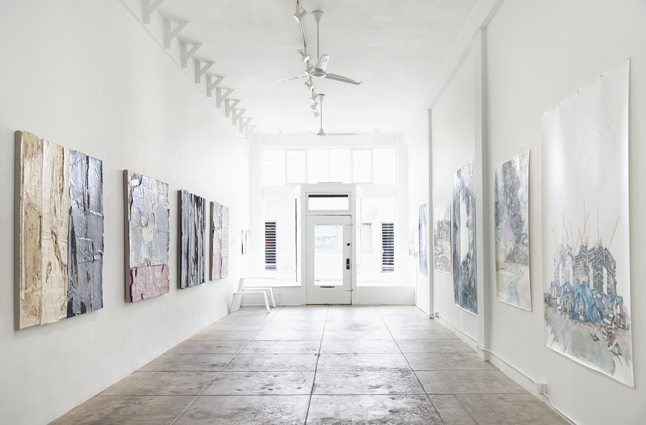 Gallery with paintings on white walls, wooden floor, and glass door at the end, illuminated by natural light.