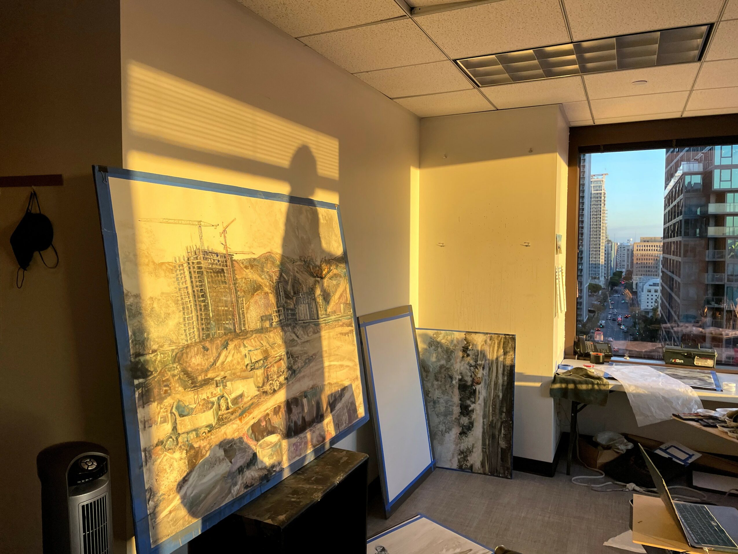 Sunlight casts shadows on a wall with framed pictures leaning against it, in an office with a window view of city buildings.