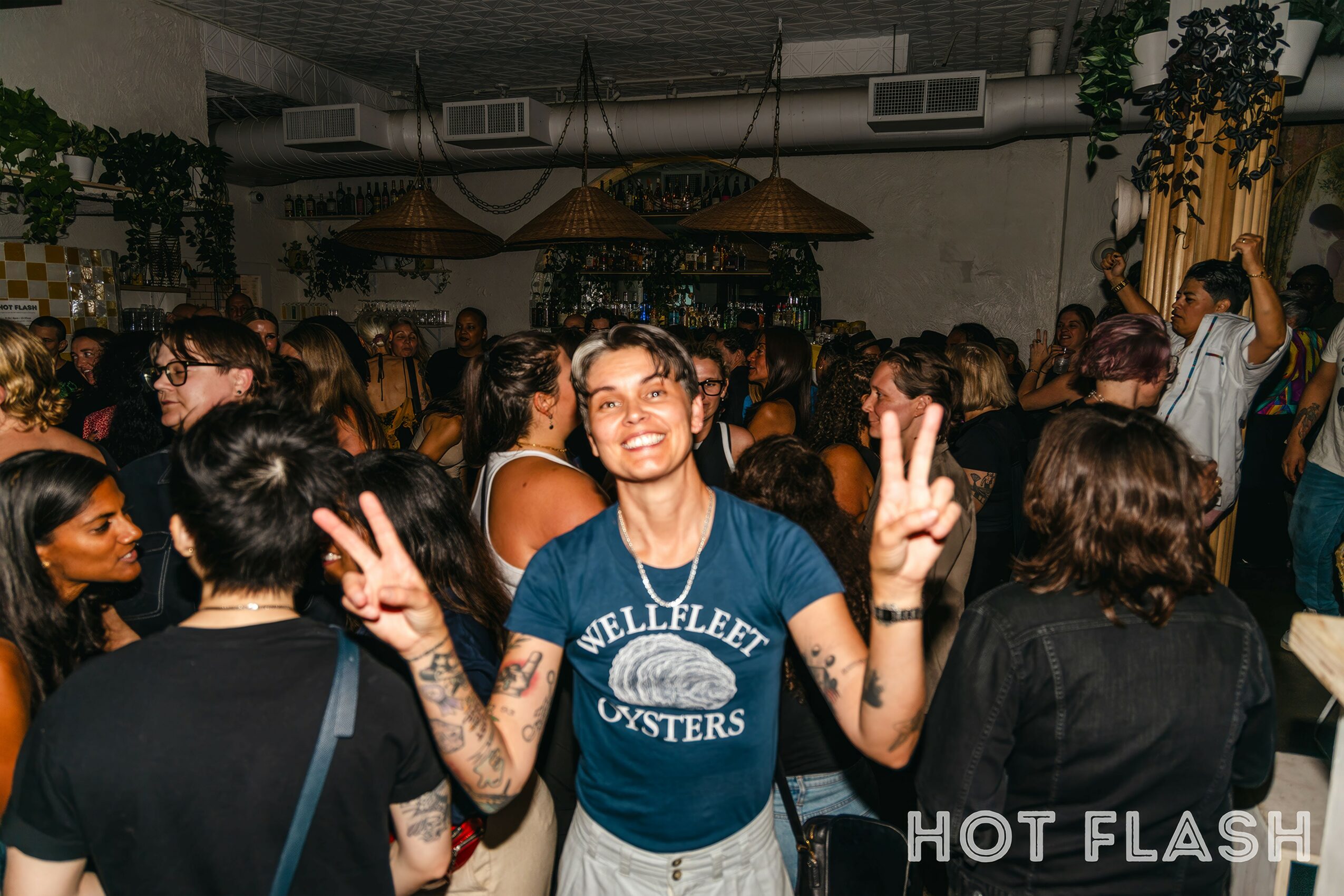 Smiling woman with tattoos making peace signs at a crowded indoor party.