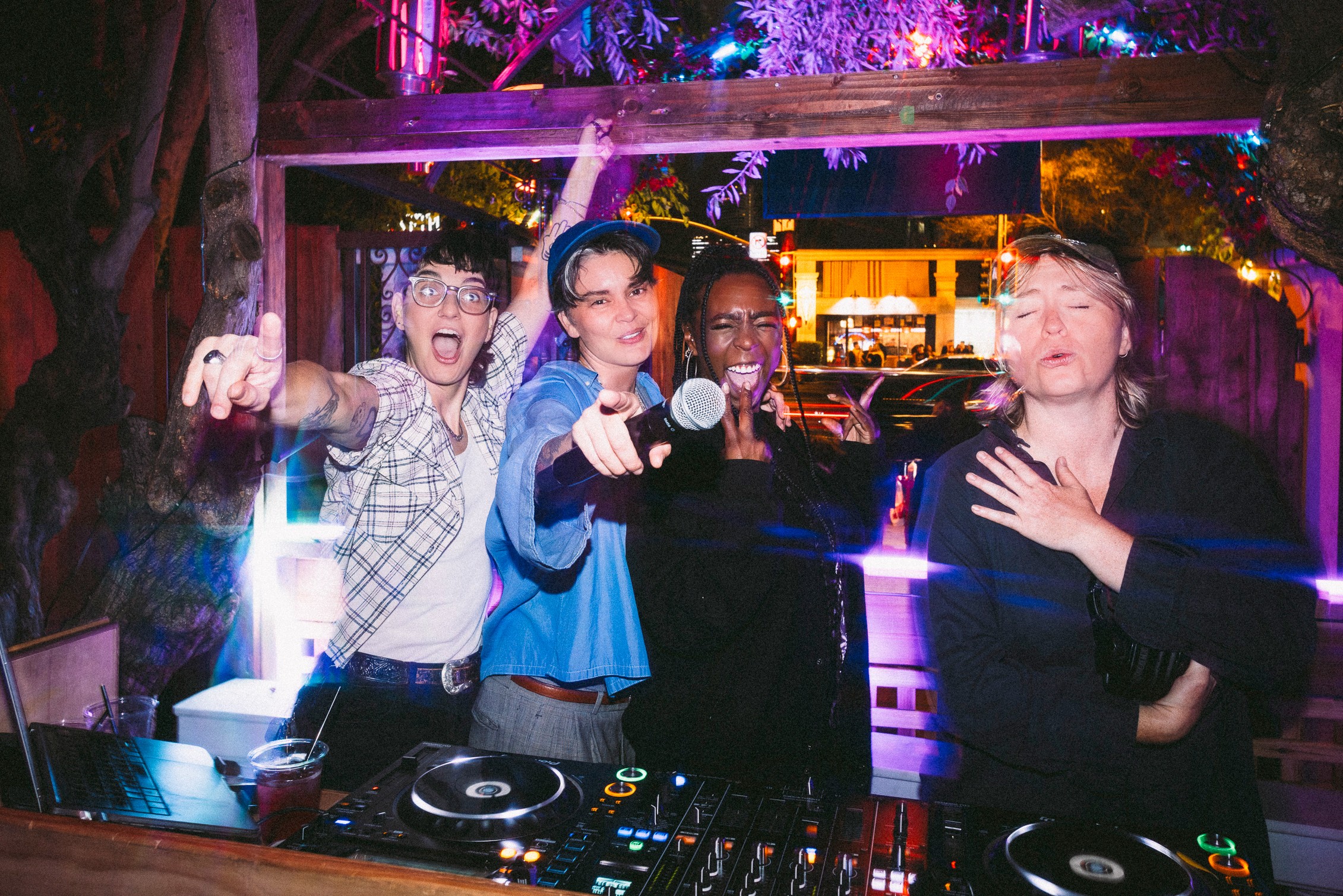 Four people at a party, smiling and pointing at the camera, with DJ equipment in front of them.