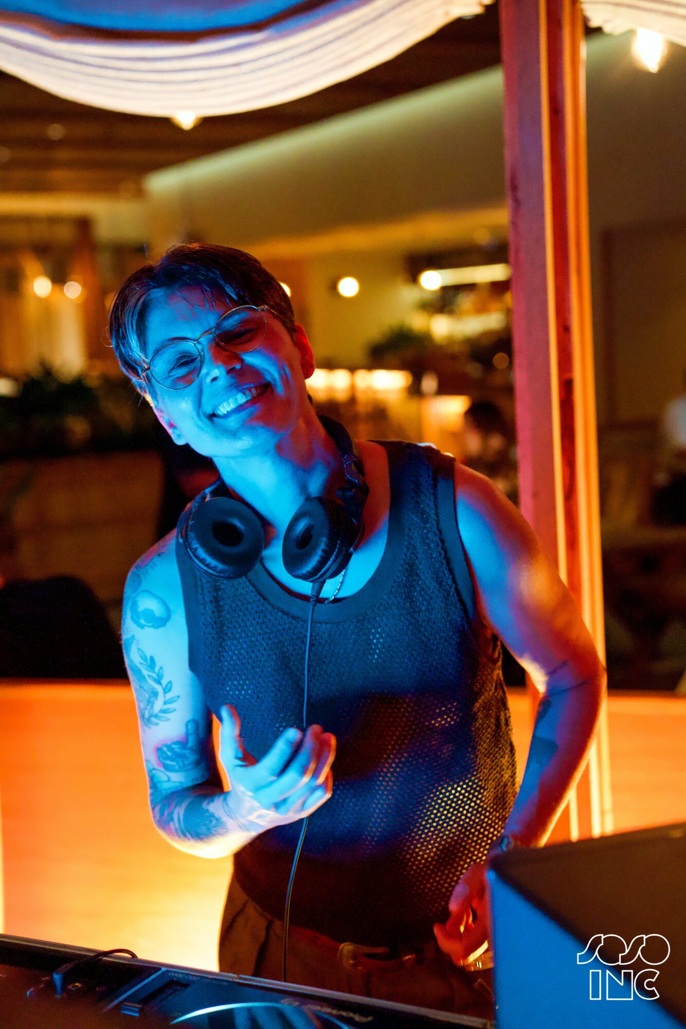 Woman with tattoos and headphones smiling at camera in colorful indoor setting.
