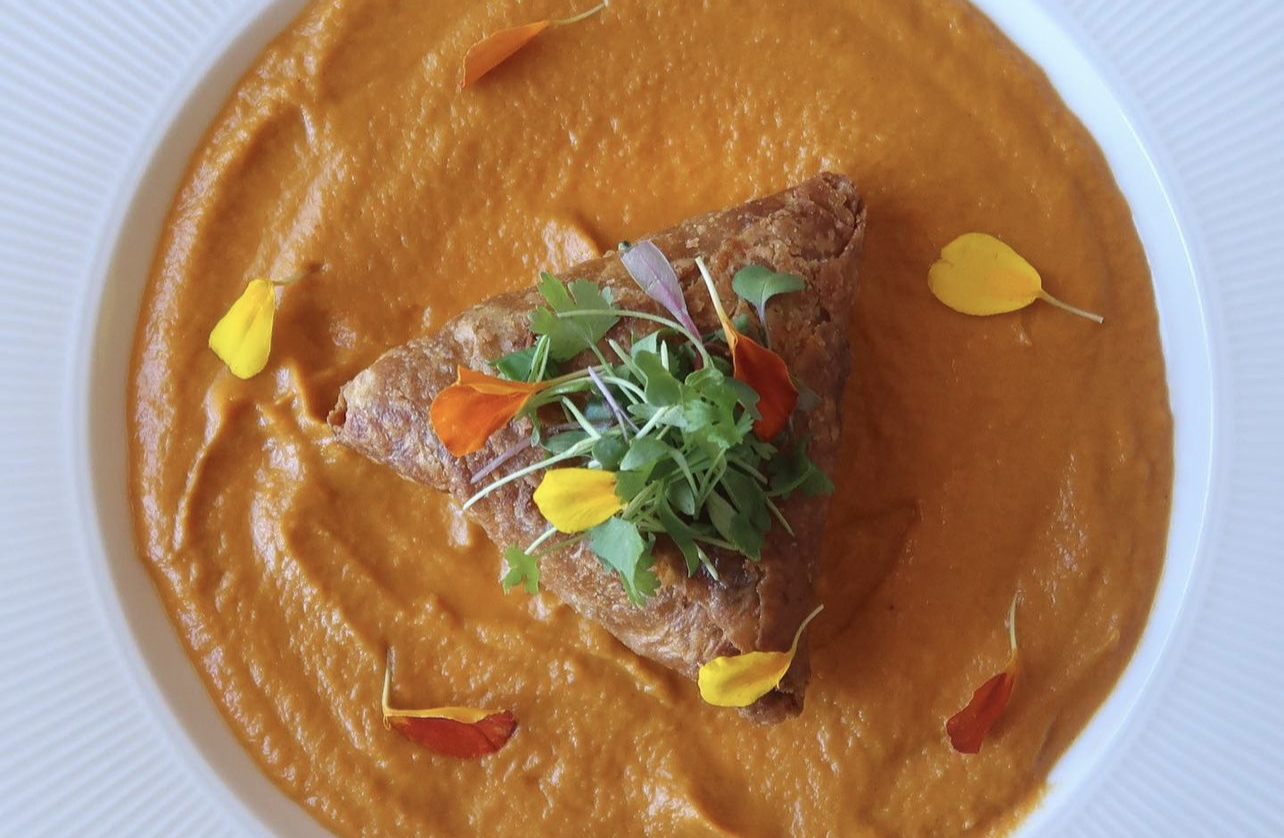 Plate with orange-colored puree, topped with a piece of meat, greens, and colorful flower petals.