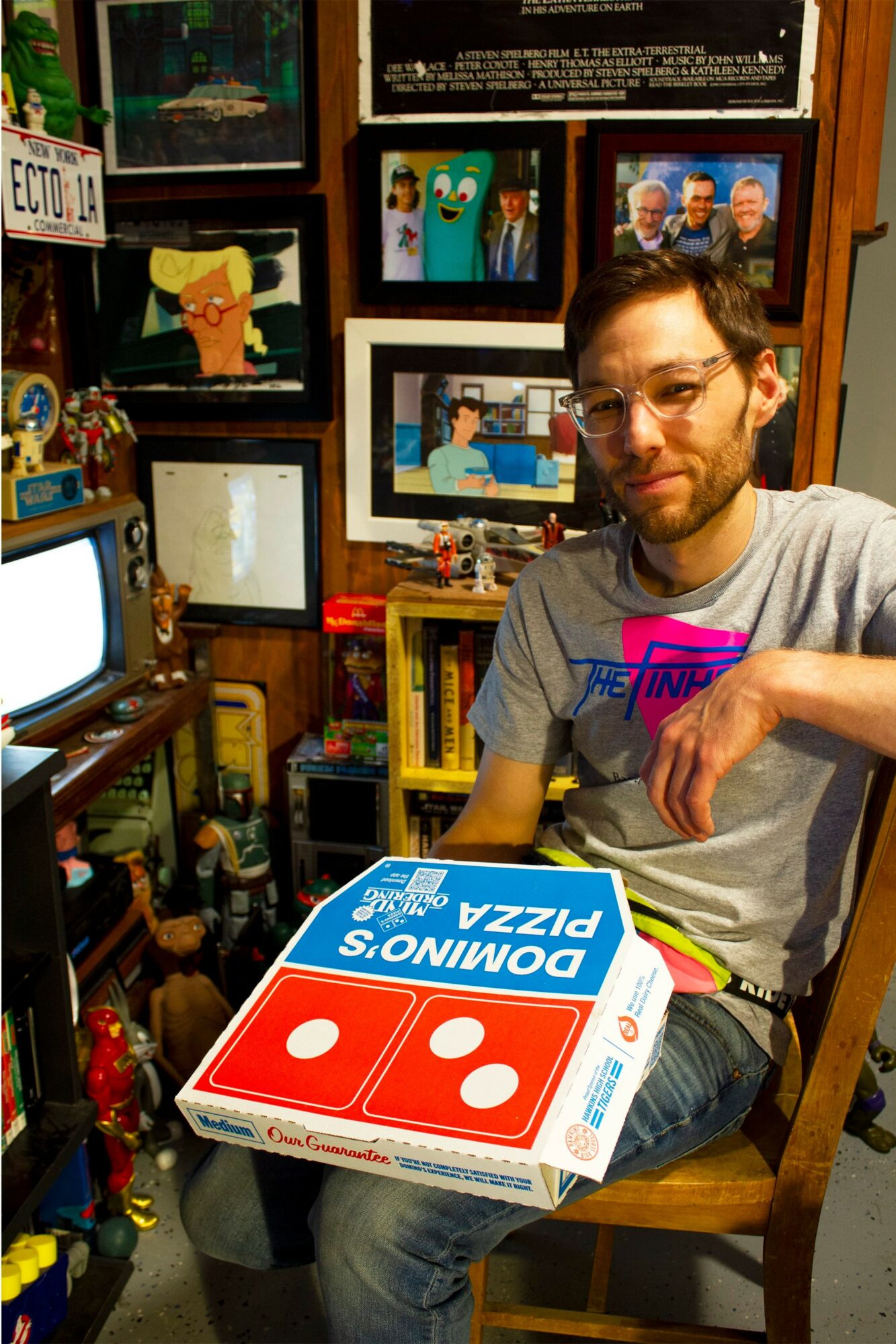 Man with glasses sitting in a room filled with framed pictures and shelves, holding a Domino's pizza box.