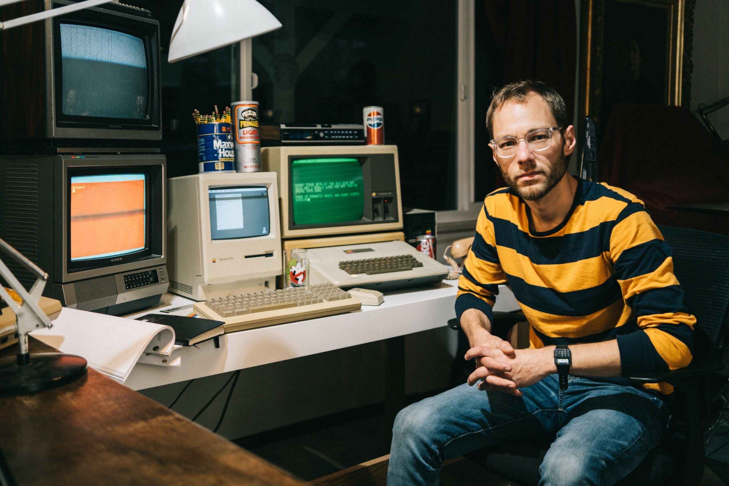 Man with glasses and beard wearing a yellow and black striped shirt sitting at a desk with vintage computer monitors and equipment.