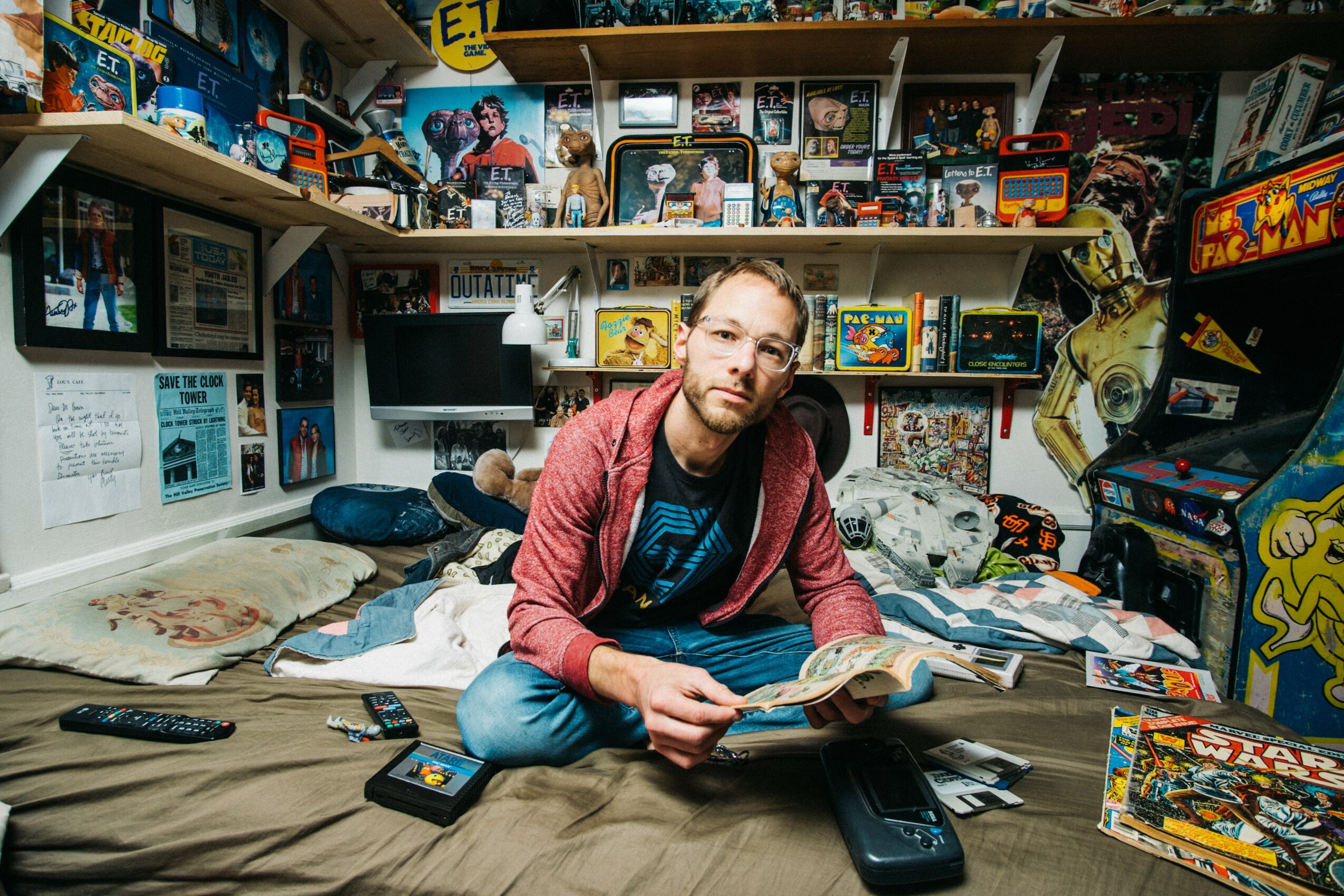 Man with glasses and beard sitting on bed surrounded by comic books and toys in a cluttered room.