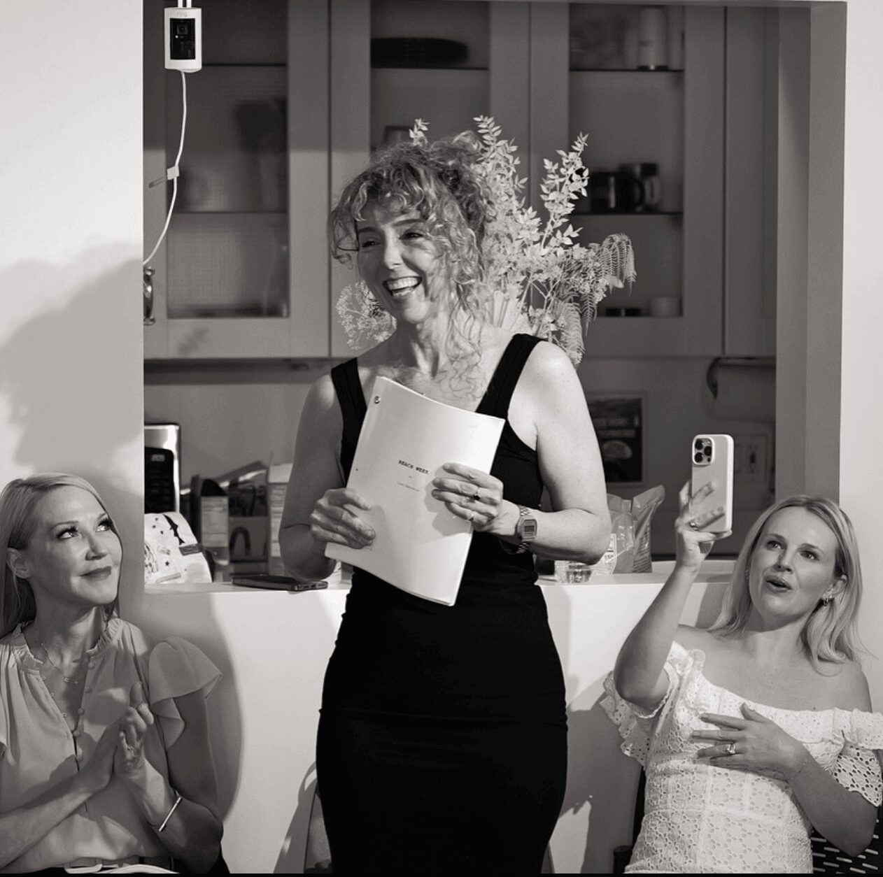 Woman standing and smiling, holding papers, with two women sitting and taking photos in a kitchen.
