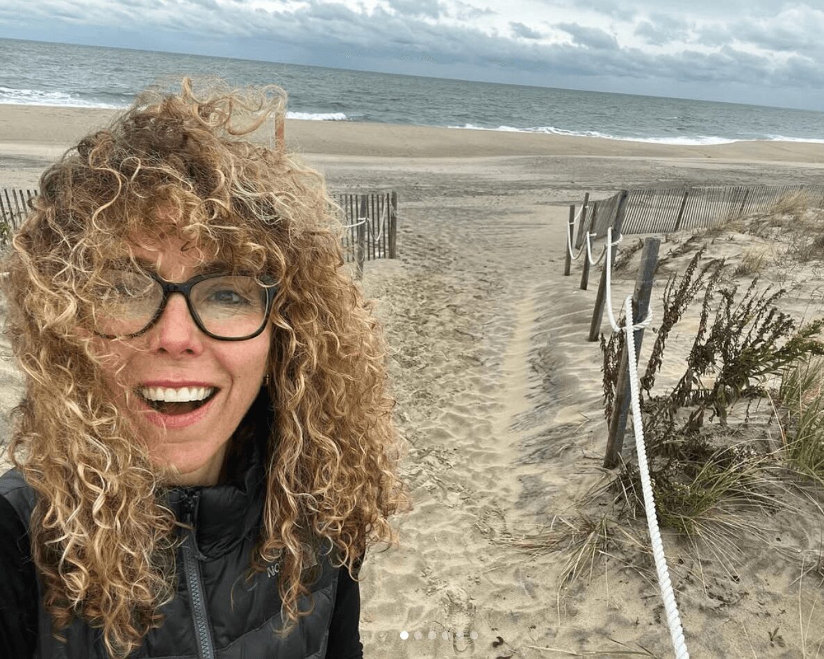 Woman with curly hair and glasses smiling at the beach, sandy path and ocean in background.
