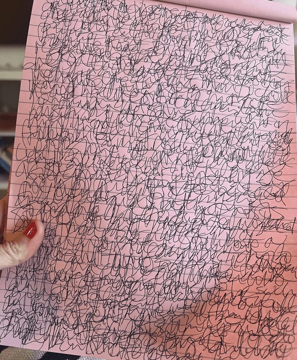 Hand holding pink notebook with densely scribbled black lines, covering entire page.