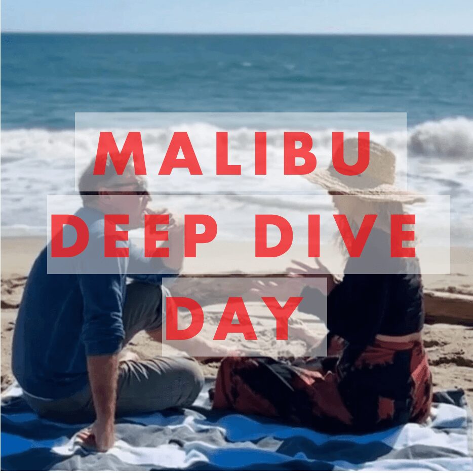 Two people sitting on a beach, facing the ocean, with a sign reading 'Malibu Deep Dive Day' in front of them.