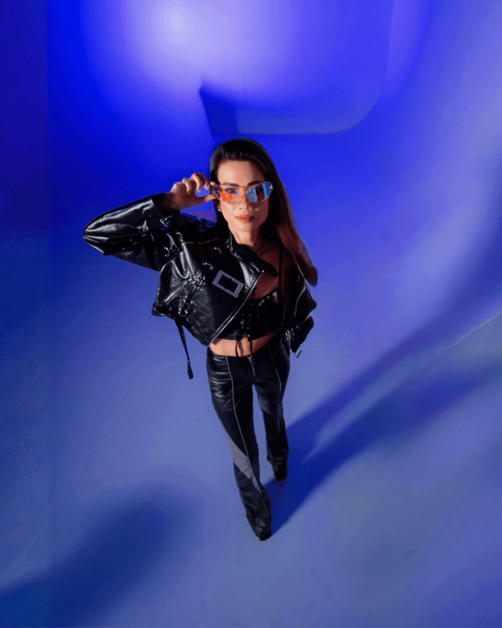 Woman in black leather jacket and pants, wearing glasses, standing in a blue-lit space, looking at the camera.