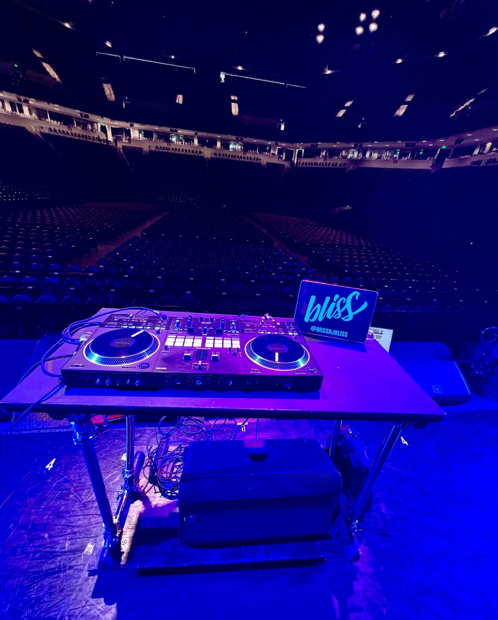 DJ equipment on a table with a laptop displaying 'Wisc' in a large venue with empty seats.