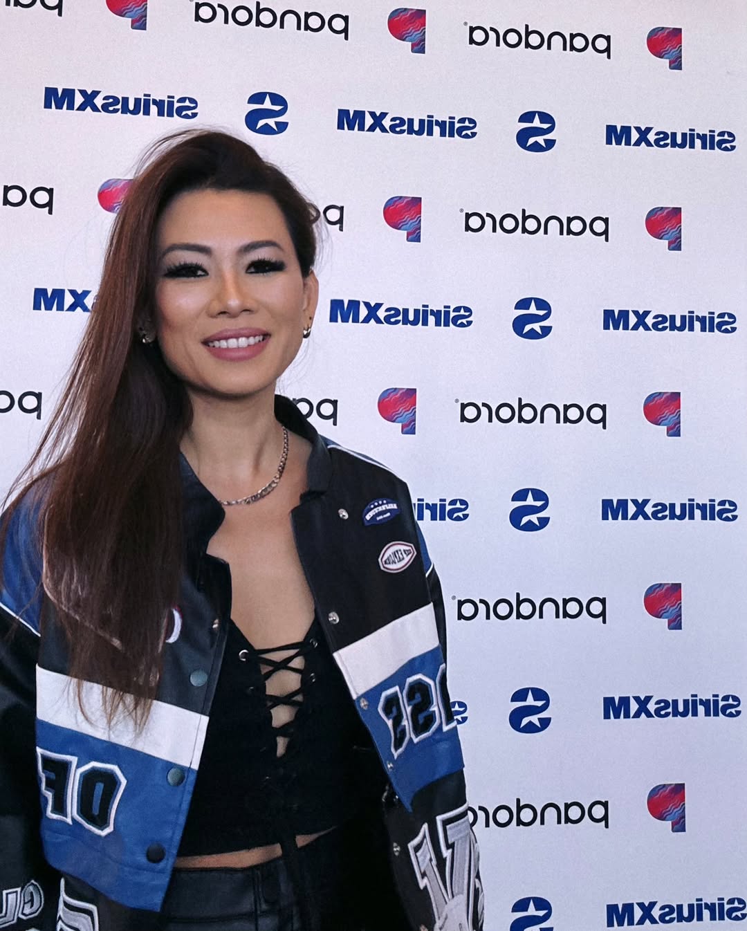 Woman with long hair smiling, wearing a black top and a blue jacket, standing in front of a branded backdrop.