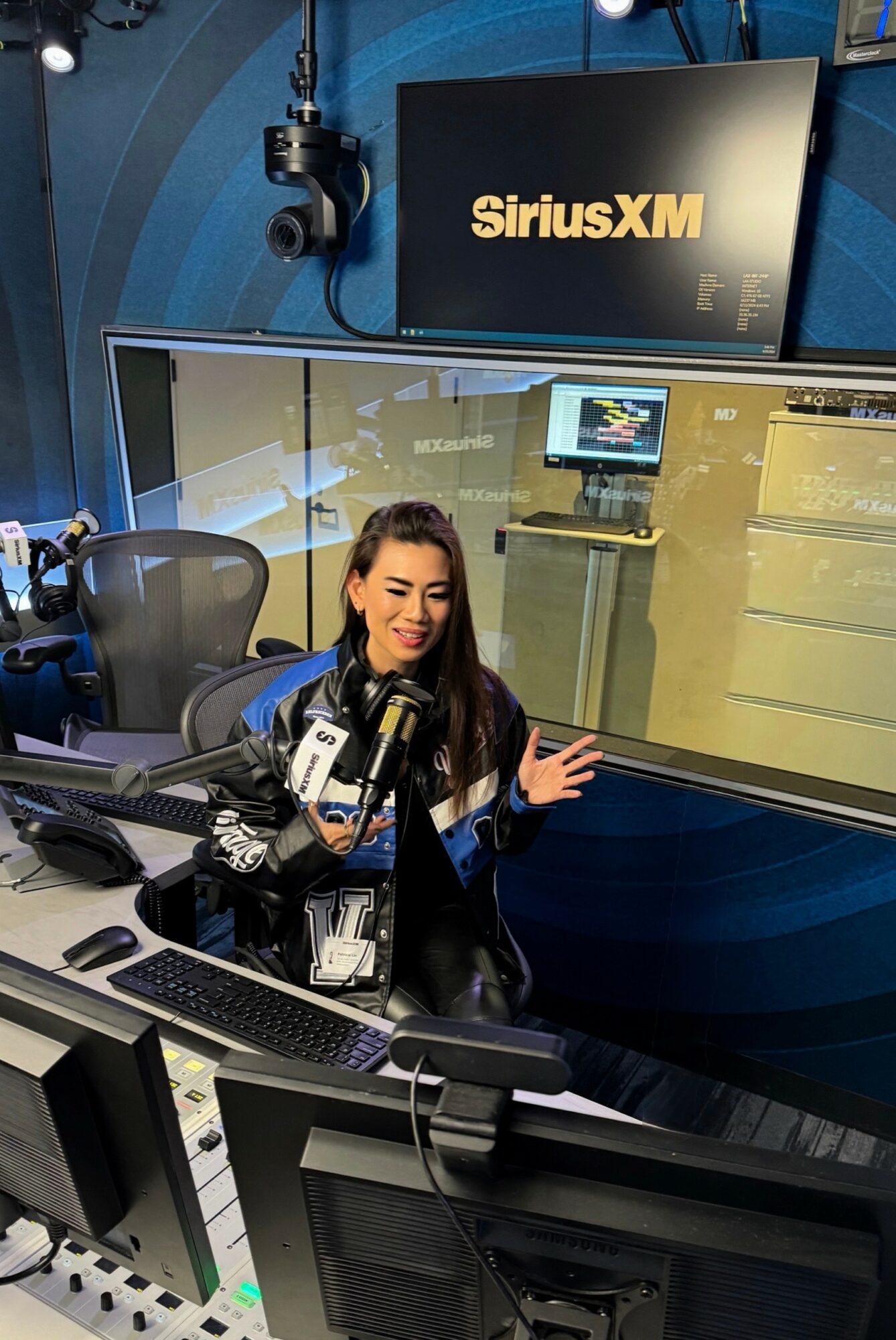 Woman sitting at a radio station desk with microphones, in front of a glass window with a SiriusXM sign.