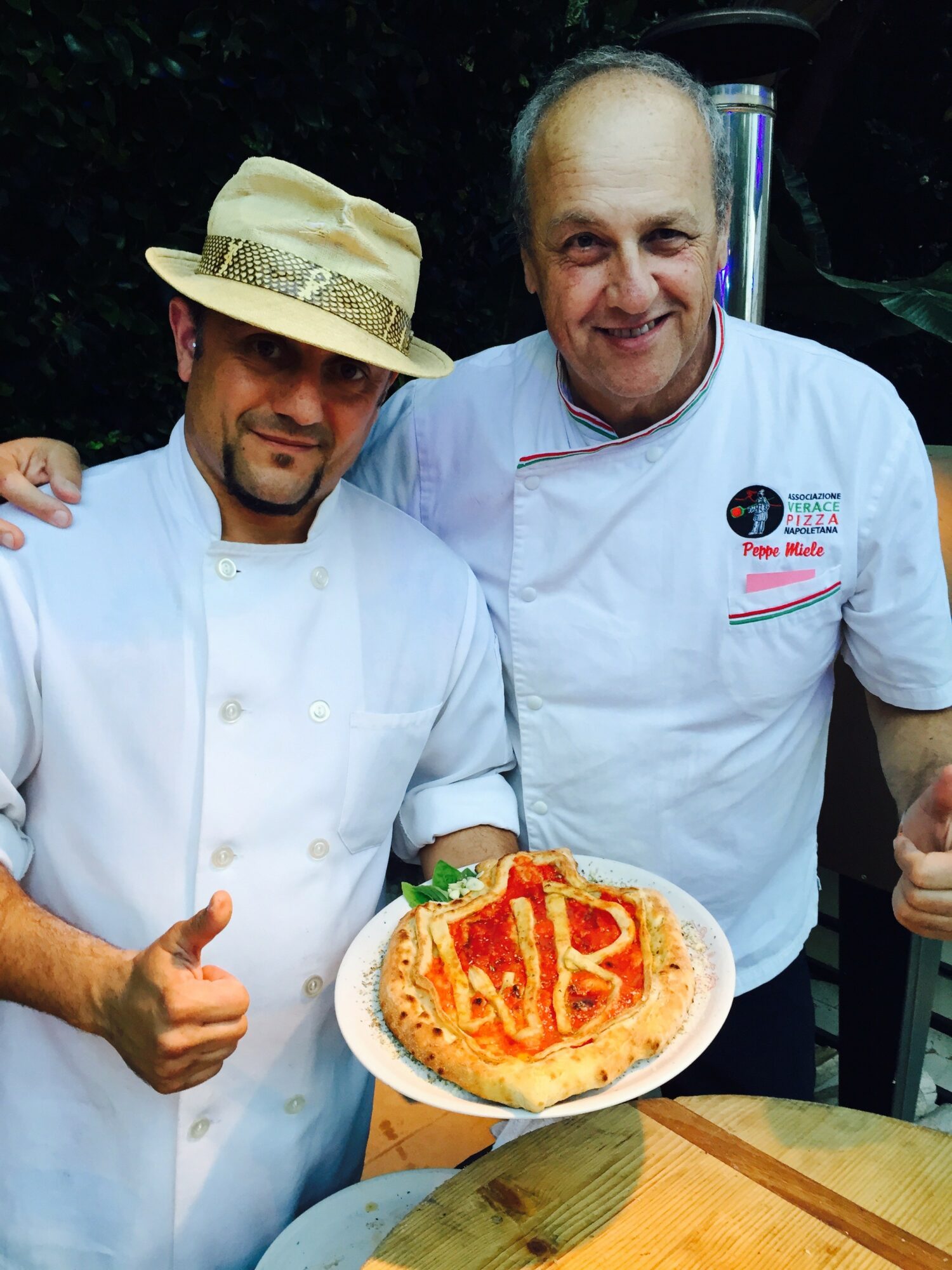 Two chefs in white uniforms holding a cake with red and yellow decorations, giving thumbs up, outdoors at night.