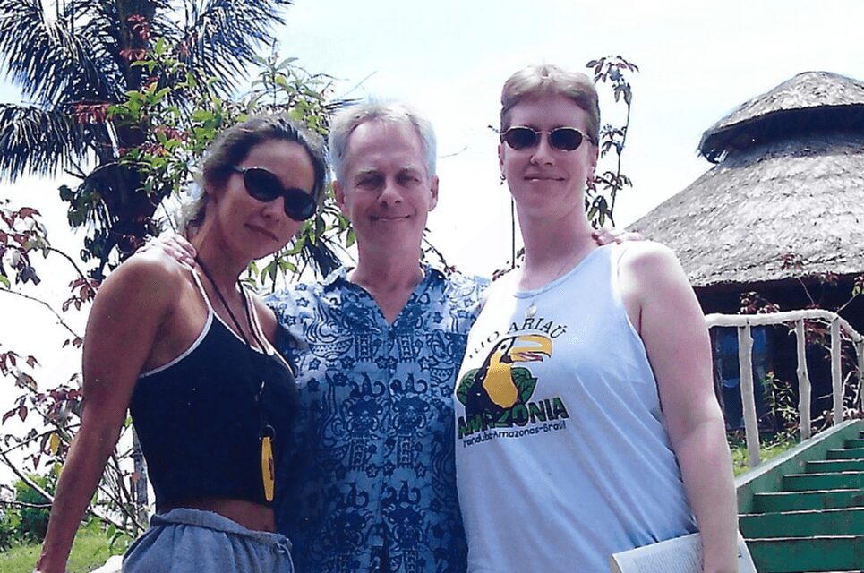 Three people standing outdoors with trees and a thatched roof structure in background.