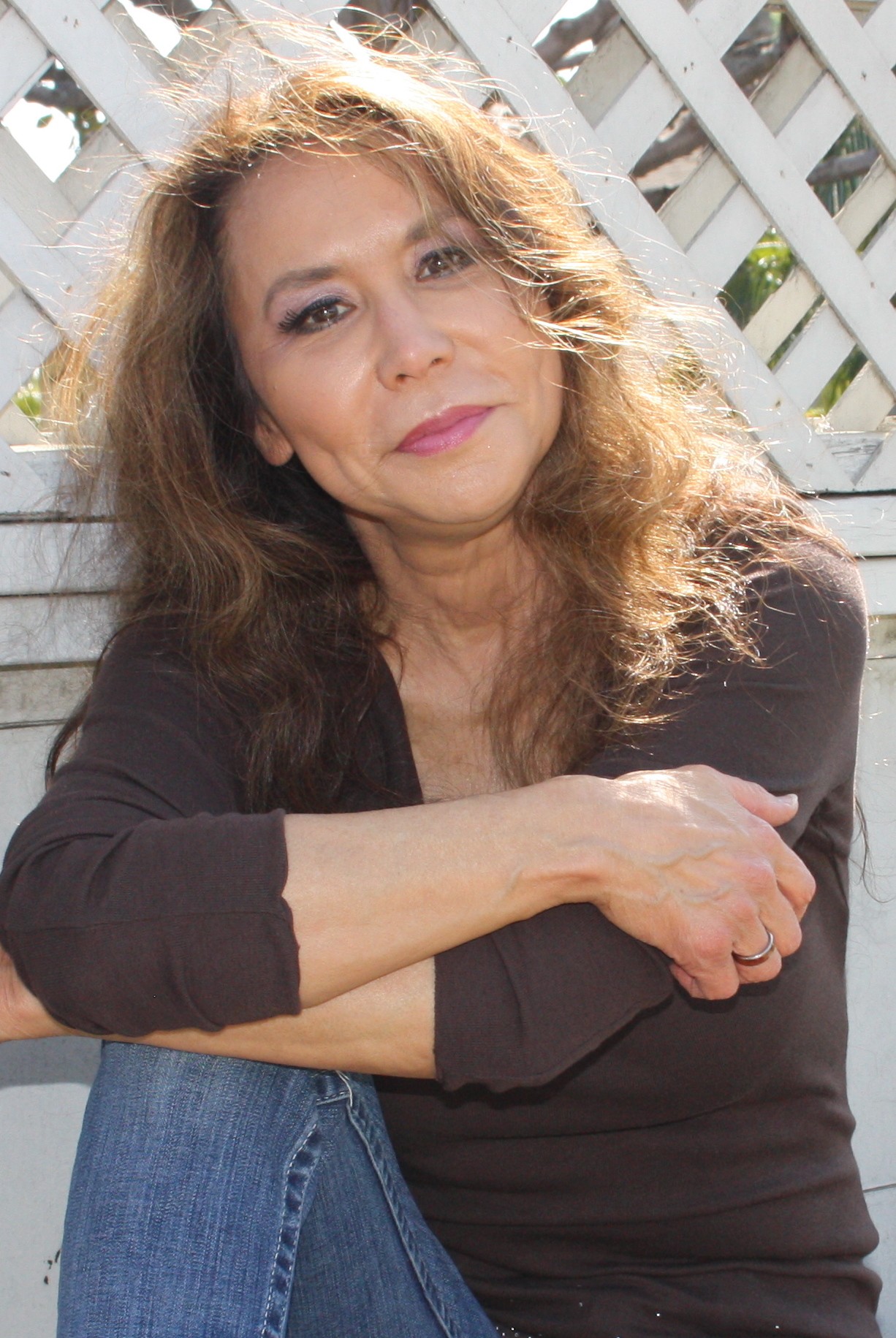 Woman with wavy hair sitting outdoors, arms crossed, looking at the camera, with a white lattice background.