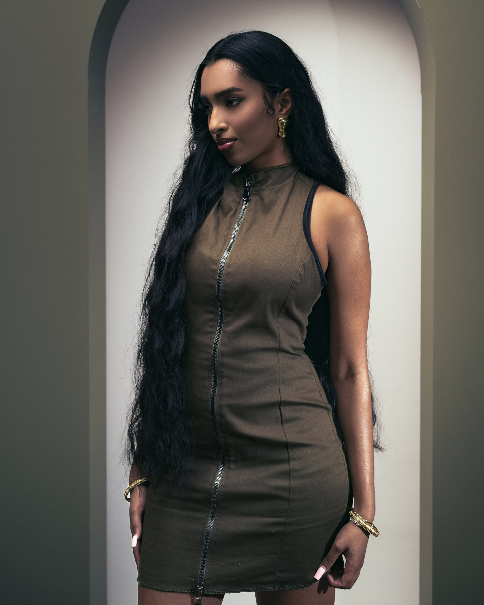 Woman with long dark hair wearing a sleeveless, zip-up dress standing in front of an archway.
