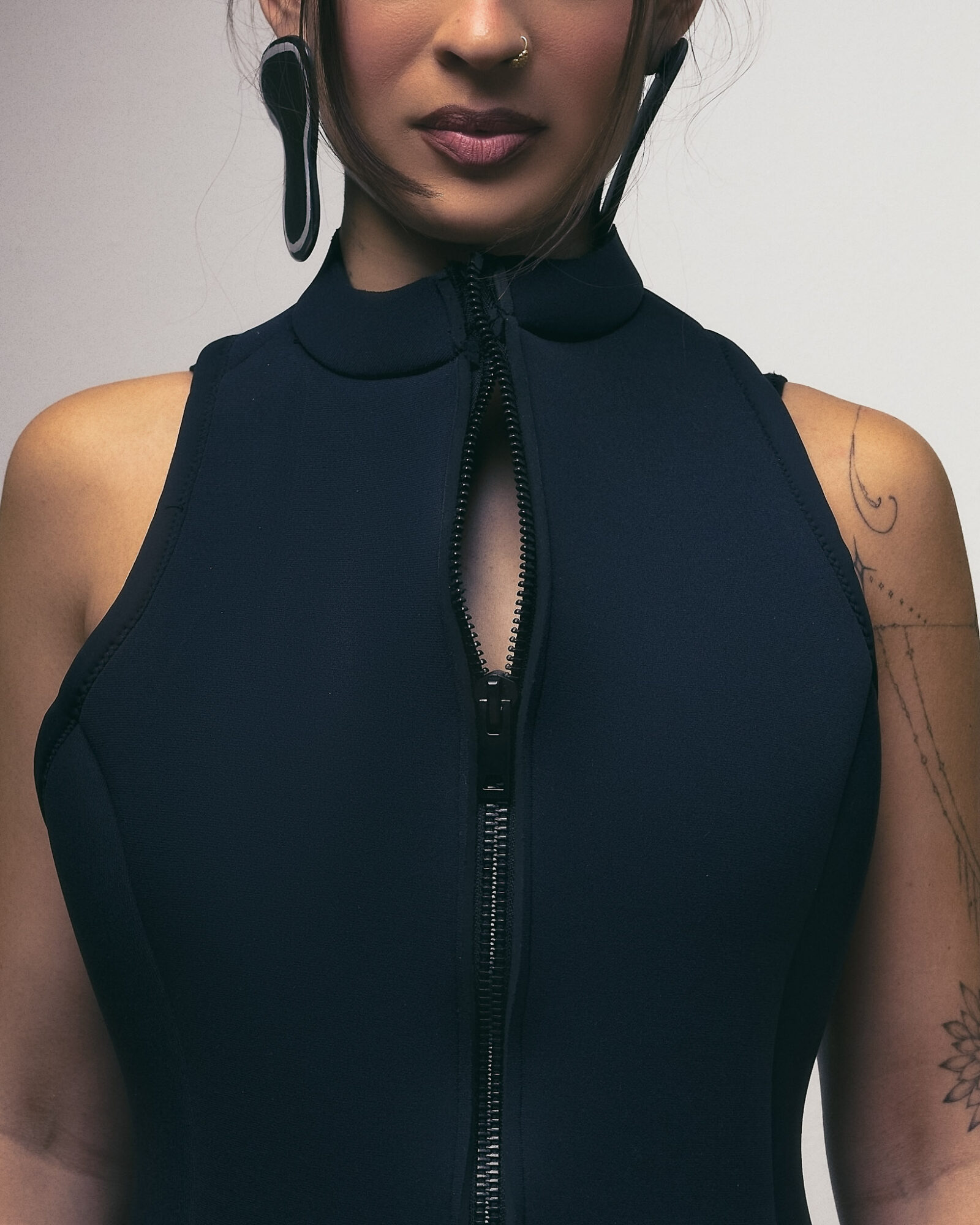 Close-up of a woman wearing a sleeveless dark blue top with a zipper, earrings, and tattoos on her arm.