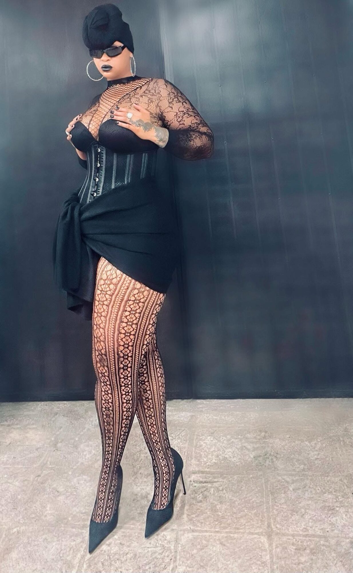 Person wearing sunglasses, lace top, corset, patterned tights, high heels, standing against dark wall.