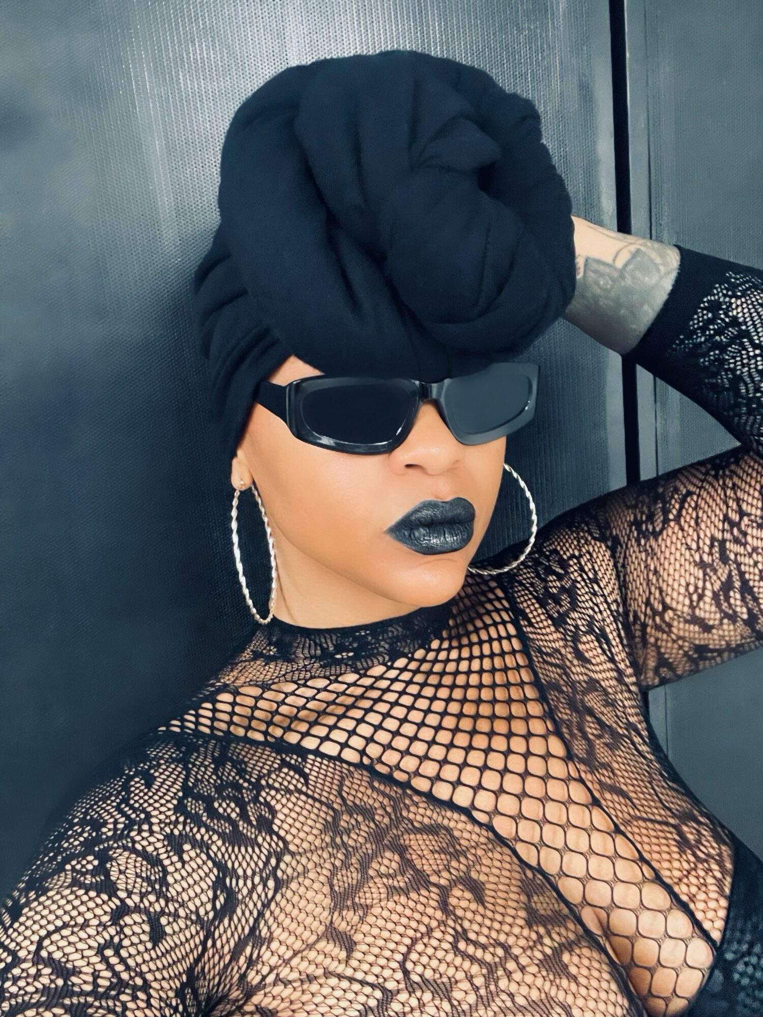 Person with black headwrap, sunglasses, dark lipstick, lace top, and hoop earrings, posing against a dark background.