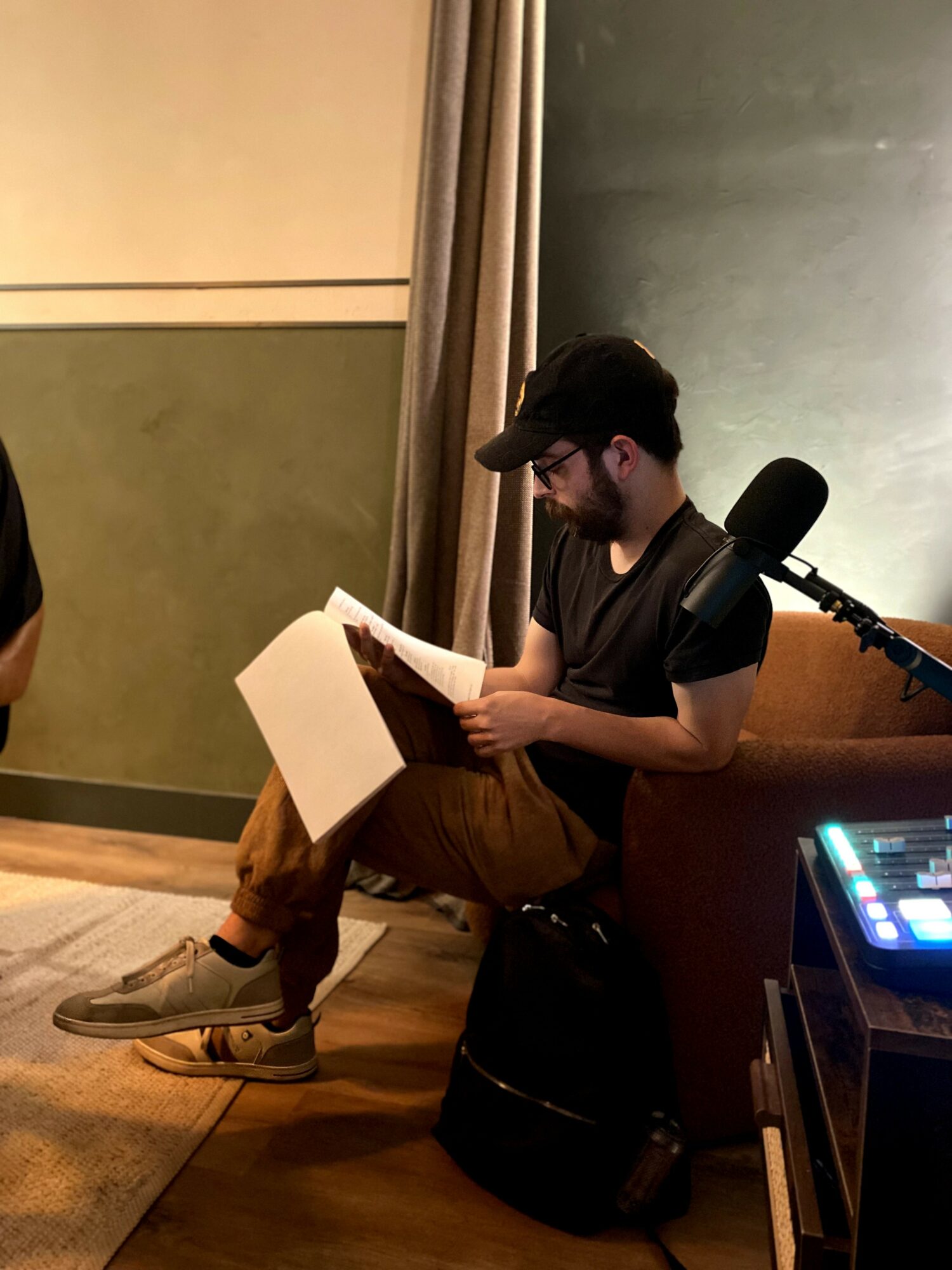 Person sitting on a stool, reading a book, with a microphone nearby in a room with green and beige walls.