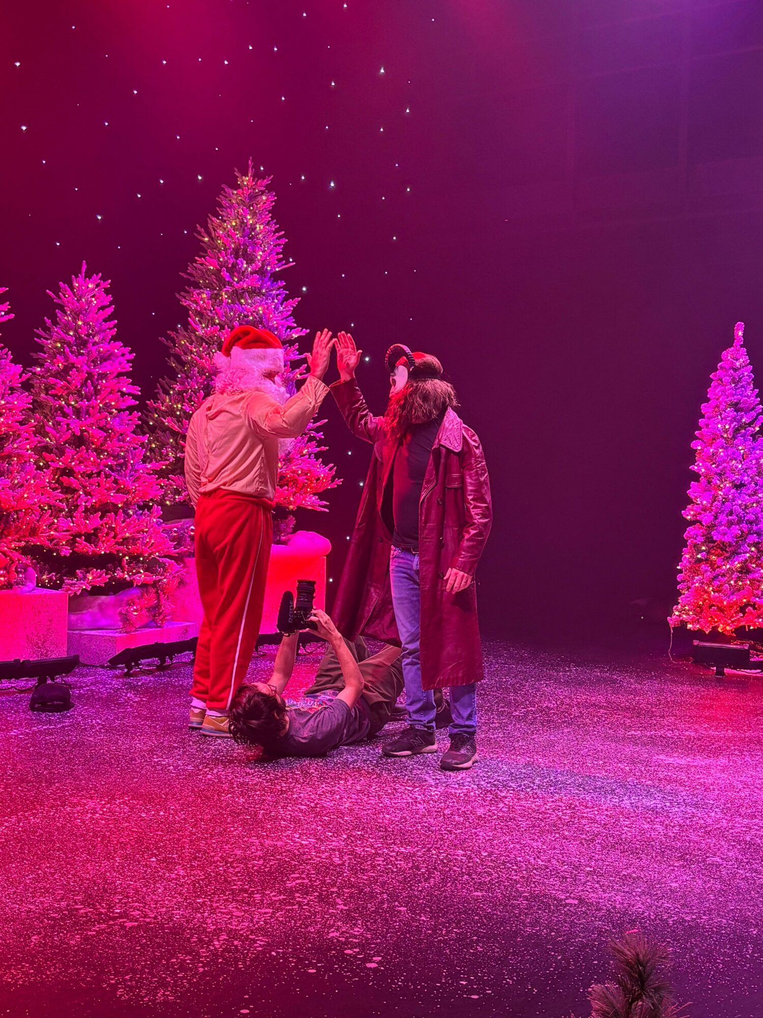Group of five people on stage with decorated Christmas trees, pink and purple lighting, some standing, one lying on the floor.