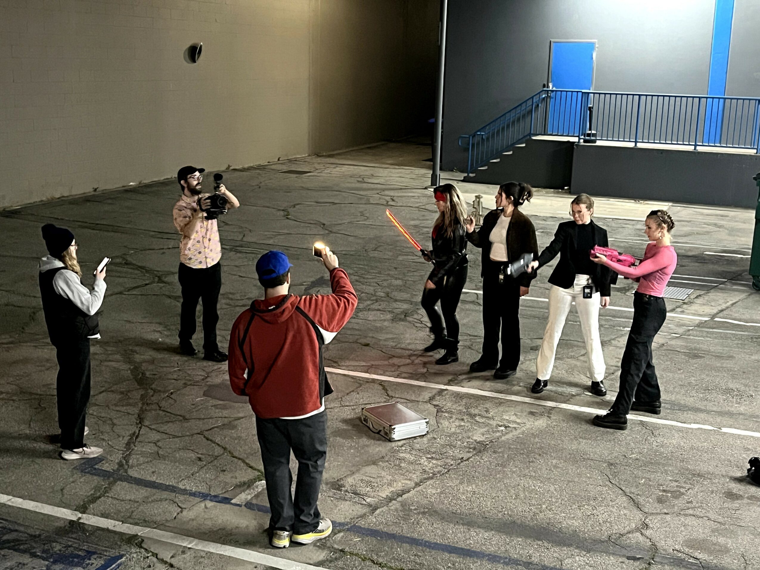 Group of seven people standing in a parking lot, some holding objects, with a person taking a photo.
