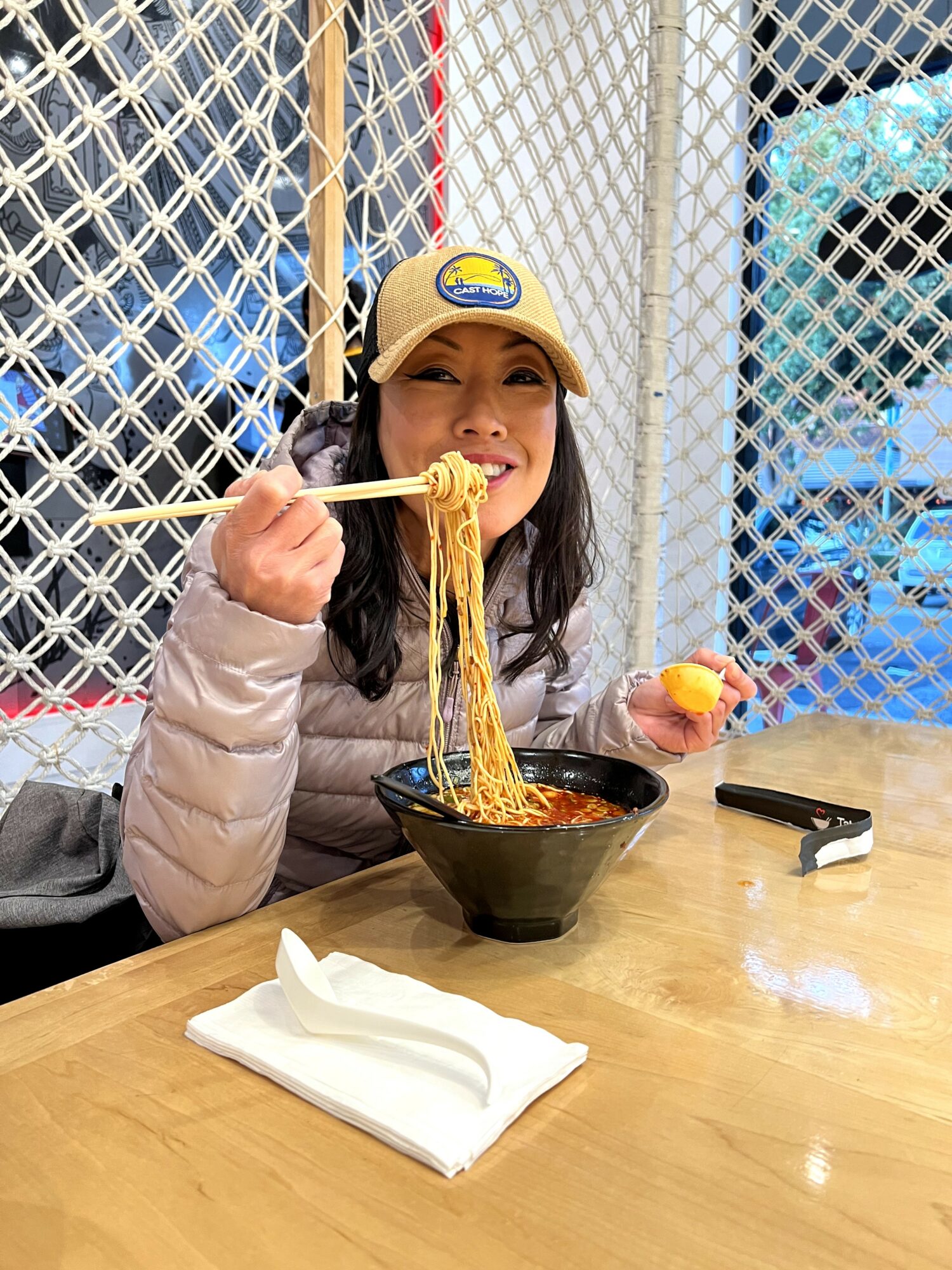 Woman wearing a yellow cap eating noodles with chopsticks at a table, holding an orange fruit in her other hand.