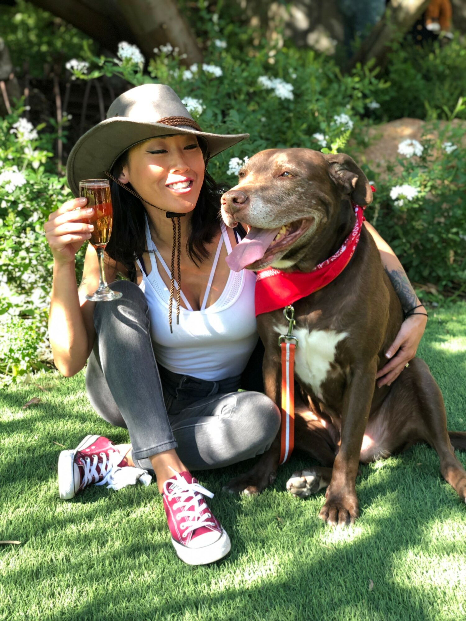 Woman with a hat and sunglasses sitting on grass, holding a glass, with a large dog wearing a red bandana beside her.