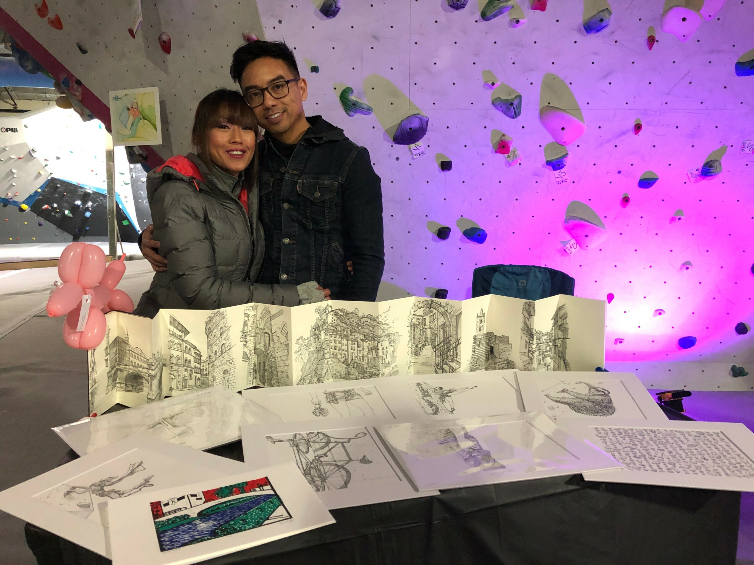 Two people stand behind a table with sketches and papers, smiling, in front of a purple-lit climbing wall.