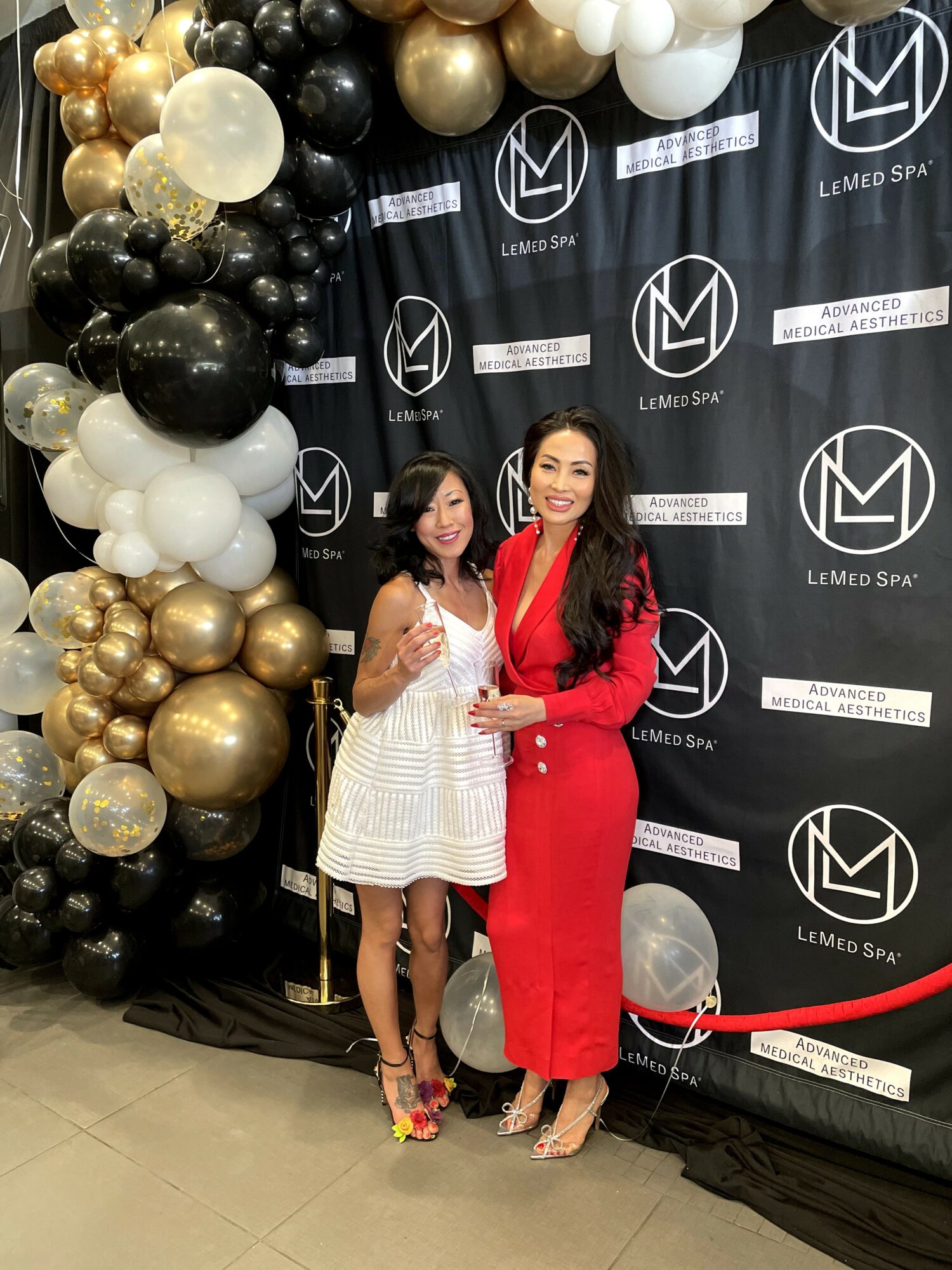 Two women stand in front of a black backdrop with balloons and logos, smiling and holding a glass award.