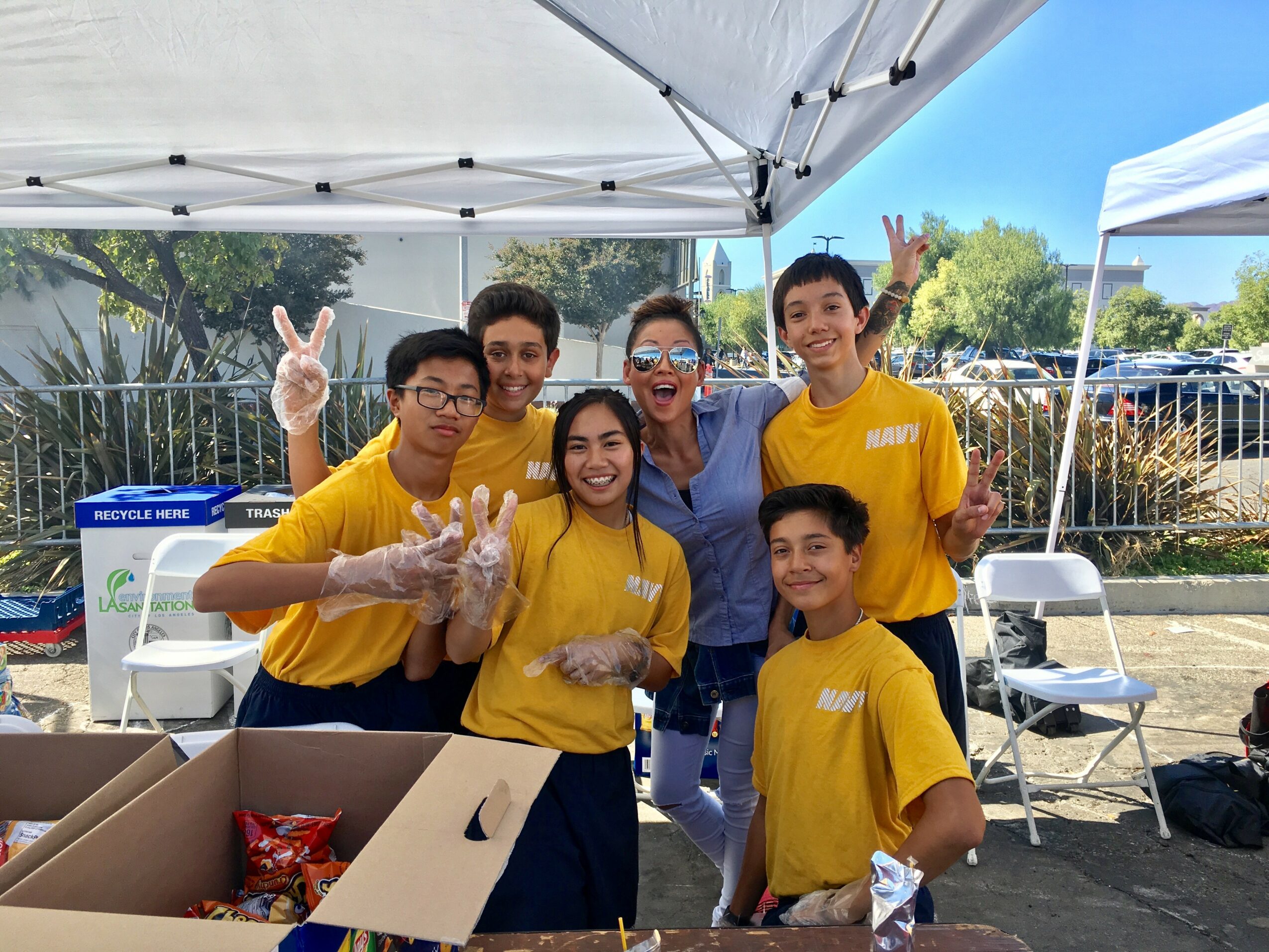 Group of six people smiling and making peace signs under a canopy, some wearing yellow shirts, outdoors.