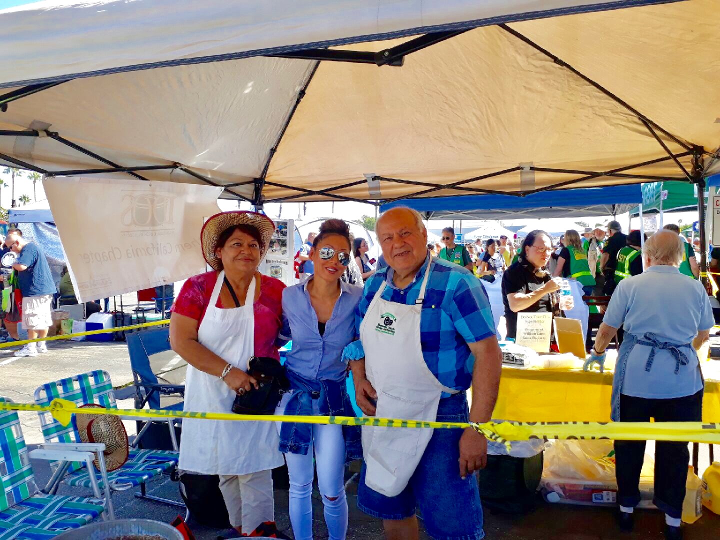 Three people standing under a tent at an outdoor event, with tables and other people in the background.