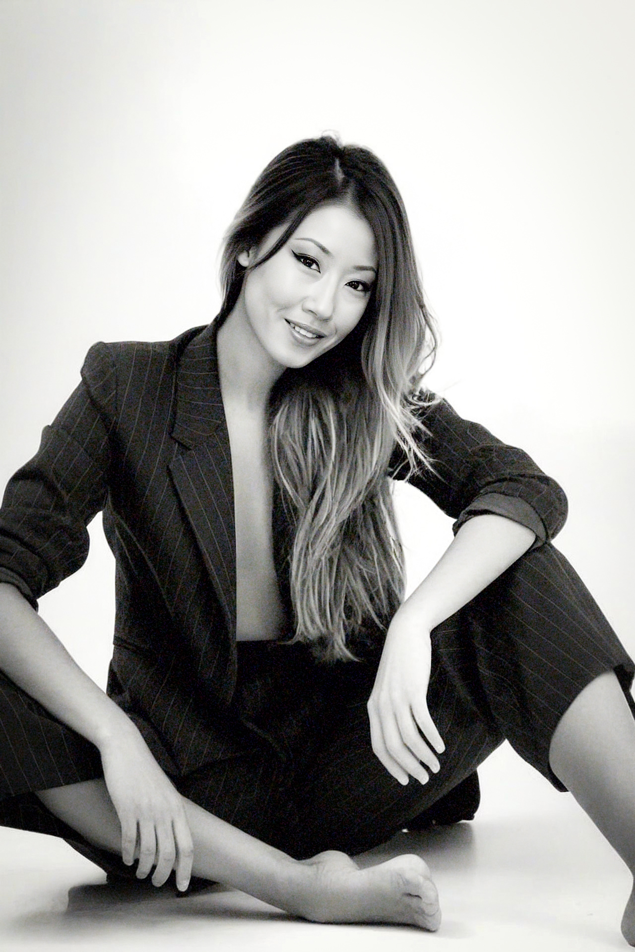 Woman with long hair sitting on the floor, smiling, wearing a dark blazer and pants, against a plain background.