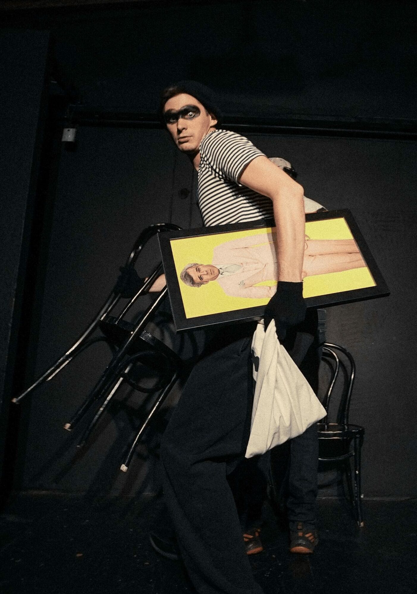 Person with glasses holding a framed photo, wearing striped shirt and black pants, standing in a dark room.