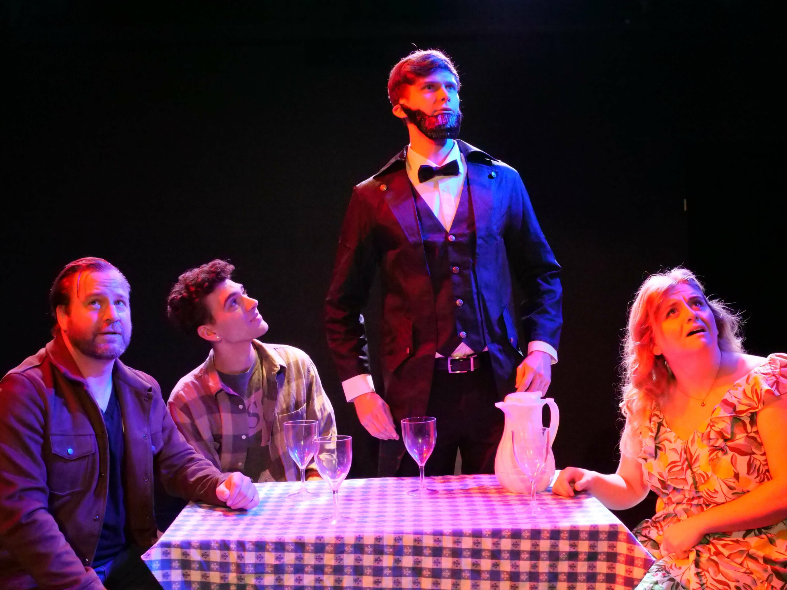 Four people at a table with a checkered tablecloth, one standing, three seated, in a dark setting.