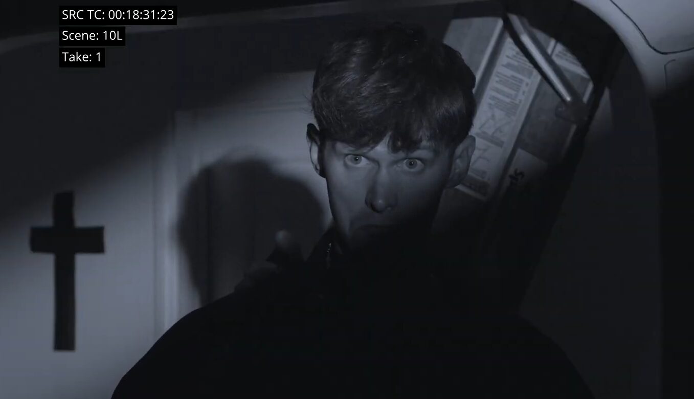 Young person with short hair looking up, shadows cast on face, dark background with a cross on the wall.