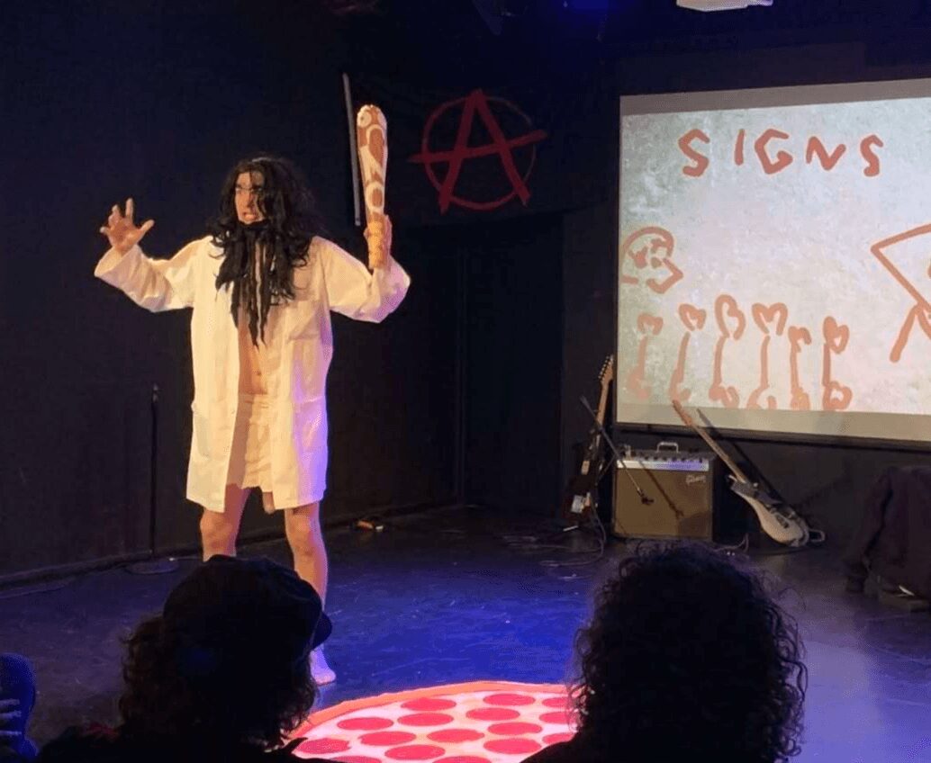 Person with long dark hair wearing a white coat on stage, gesturing with hands, audience in foreground, presentation slide visible.